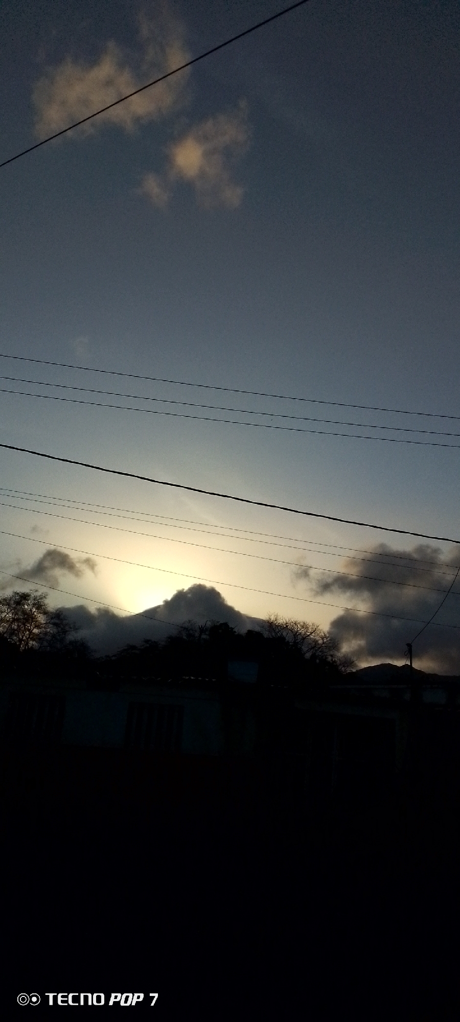 The image shows a landscape at dusk in Altagracia, Venezuela. In the middle distance, a mountain range is silhouetted against a muted yellow and gray sky. Clouds partially obscure the peaks. Power lines crisscross the upper half of the frame. In the foreground, a dark, indistinct shape suggests buildings and trees. The lighting is dim, emphasizing the contrast between the bright sky and the dark foreground. The bottom of the image includes the phone's model.