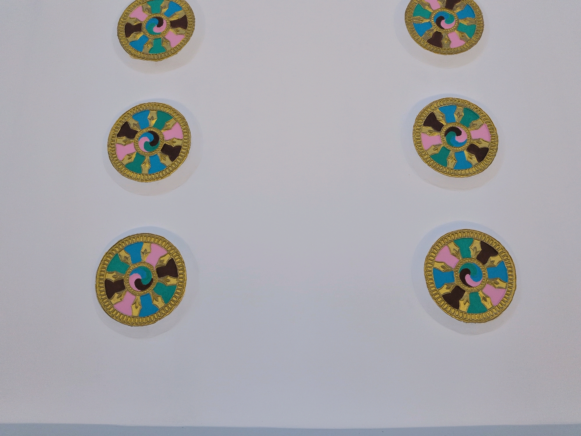 Six circular decorative medallions with intricate gold borders are arranged in two vertical columns on a plain white wall. Each medallion features a spiral design in the center, with alternating segments of turquoise, pink, brown, and a darker color, possibly black or dark brown. The medallions are evenly spaced, with three in the left column and three in the right column, positioned at different heights. The background is a smooth, white surface, possibly a wall or ceiling. The image has a shallow depth of field, with the medallions in focus and the background slightly blurred. There are no people or other objects visible in the scene. The style of the medallions suggests a decorative element common in Buddhist architecture or art, which is prevalent in Leh, India.