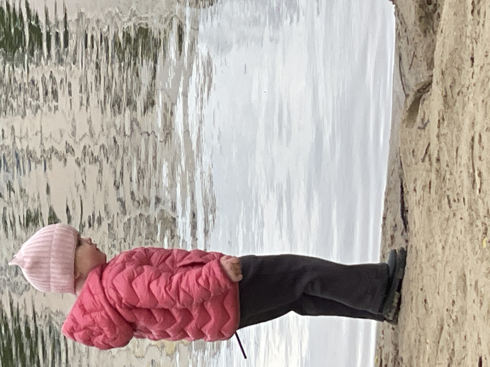 A young child wearing a pink hat and puffy pink coat, along with dark pants and boots, stands near a body of water, possibly a lake or river. The water is reflecting light and creating a shimmering effect. To the right of the child is a large rock or cliff face. The child appears to be standing next to a body of water in a park or natural area. It is difficult to verify its location without more context. Based only on the photograph, there is no way to confirm the location in the city of Moscow, Russia.