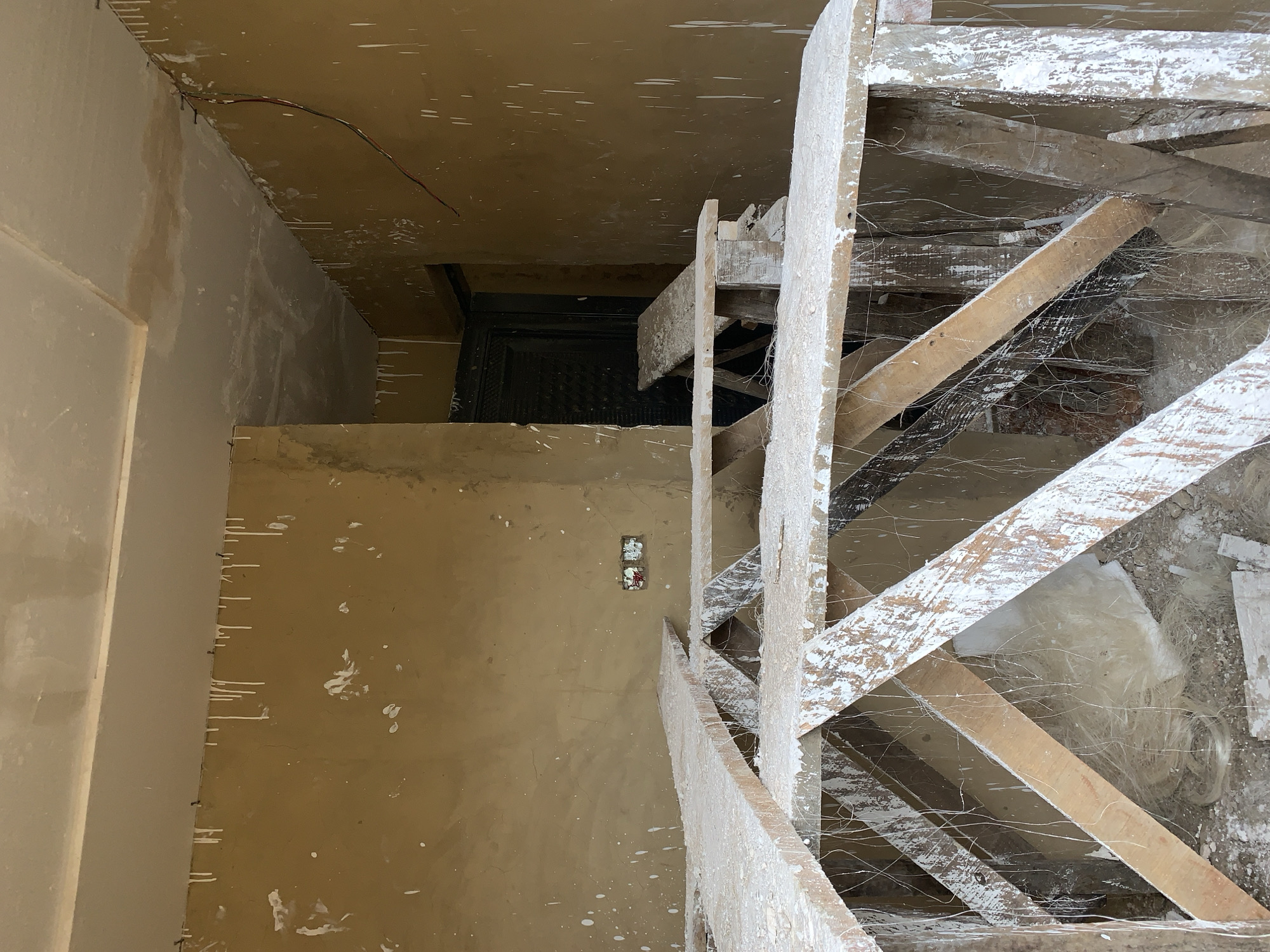 The image depicts an unfinished interior space, likely a room or hallway, located in the City of New Stadium, Nigeria. The walls are coated in a matte, light brown or tan finish and are extensively splattered with white paint. Numerous long, vertical drips of white paint run down the walls from the ceiling line, indicating recent or ongoing painting. The ceiling is white, appearing to be a plaster or drywall surface, with some areas showing slight discoloration or unfinished patches.
In the foreground and midground, several rudimentary wooden work platforms or scaffolding structures are present. These are constructed from rough-hewn timber planks and supports, heavily coated in dried white paint splatters and draped with a significant amount of fine, white, cobweb-like fibrous material. Beneath the platforms, the floor is covered in construction debris, dust, and fragments of white material.
On the left wall, two unfinished electrical outlet boxes with exposed wiring are visible. Towards the center-right of the image, in the background, a dark, possibly metal, patterned door is visible within an open doorway. On the right-hand wall, near the ceiling, a pair of red and black electrical wires hang loosely. No individuals are present in the scene. The overall environment suggests an active construction or renovation site in an incomplete state.