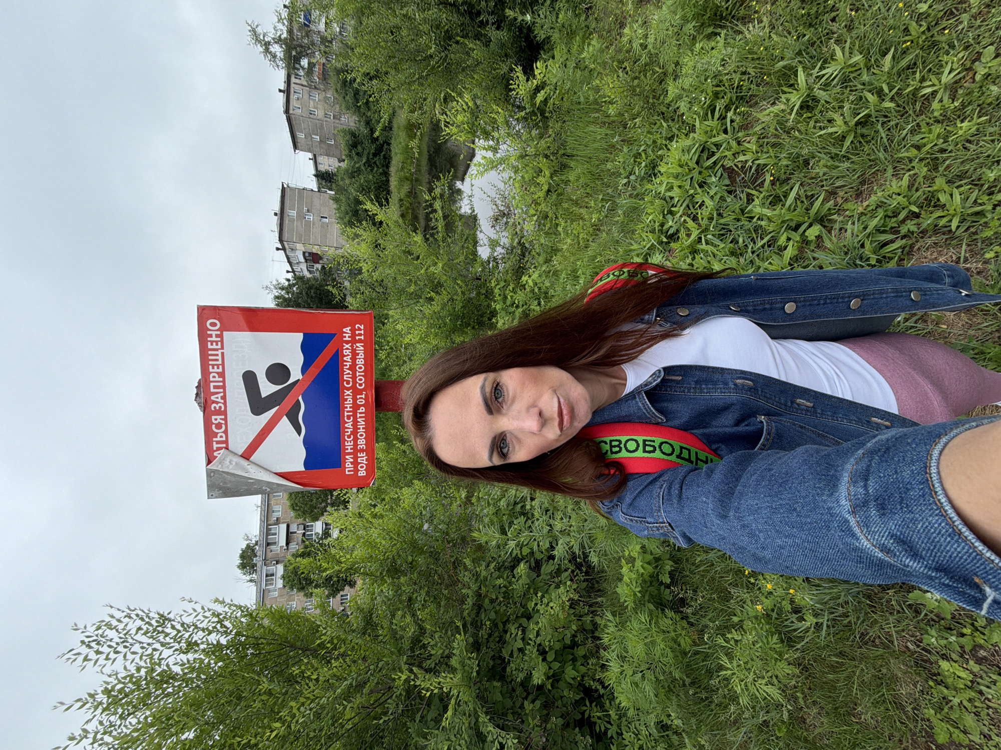 A woman with long brown hair, wearing a denim jacket, a white t-shirt, and pink leggings, takes a selfie in front of a "No Swimming" sign in Birobidzhan, Russia. The sign, with a red border and white background, features a black silhouette of a person swimming with a red diagonal line through it. Text on the sign, in Russian, advises to call 01 or 112 in case of emergencies. Behind the woman and the sign, a small body of water is visible, surrounded by lush green foliage. In the background, Soviet-era apartment buildings can be seen, typical of the city's architecture. The sky is overcast. The woman's backpack, with green lettering that reads "СВОБОДА" (Freedom), is visible.