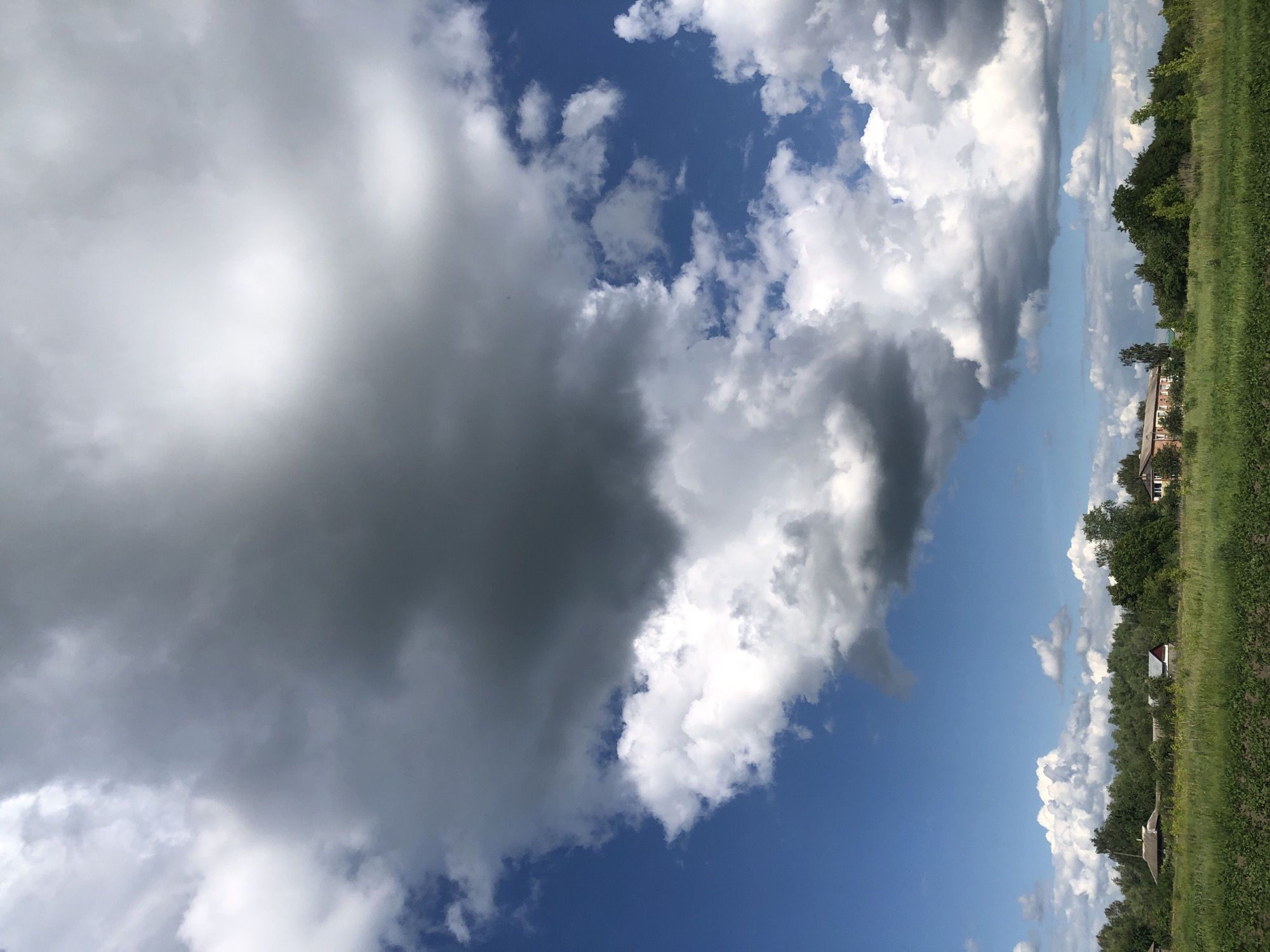 The image captures a view of the sky and a rural landscape. Dominating the upper portion of the frame are large, dramatic clouds, with some appearing dark and ominous and others bright white and puffy. The sky is a vibrant blue, with patches of lighter blue visible between the clouds. In the lower half of the image, a band of lush green grass stretches across the foreground. Beyond the grass, a line of trees forms a dense backdrop. Scattered amongst the trees are a few small buildings, some with red roofs, suggesting a small village or collection of houses. The overall impression is one of a natural landscape under a dynamic sky, possibly indicating impending weather changes. The location is identified as Khmilnytsia, Ukraine. There are no people or animals visible in the image.