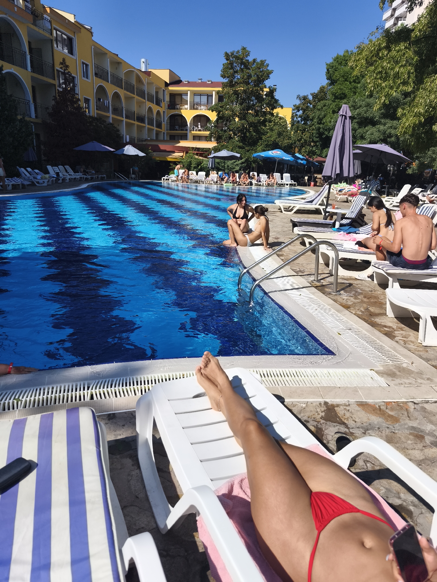 An overhead, full-shot captures a sunny day at a resort-style swimming pool. The pool, a prominent feature, has a striking checkerboard pattern of bright blue and dark blue tiles. Sunlight reflects off the water, creating ripples and a sense of depth. Around the pool, lounge chairs are arranged, some occupied by people. In the foreground, a person's legs are visible, extended on a white lounge chair with a pink towel. They are wearing a red bikini bottom. To the left, another striped lounge chair has a dark object on it.
Further back, several individuals are seen relaxing by the pool. Some are sitting on lounge chairs, while others are near the water's edge. The background reveals a large, yellow, multi-story building with arched balconies, typical of a hotel or resort. Lush green trees and foliage surround the area, and several large, closed umbrellas are strategically placed, suggesting shaded seating areas. The sky is a clear, bright blue. The overall scene depicts a leisurely and tranquil atmosphere at a vacation spot.