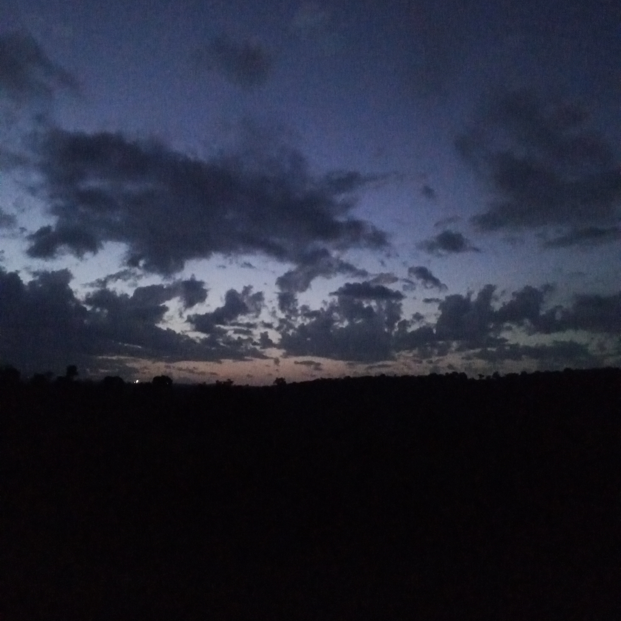 The image depicts a landscape scene at dusk or night, potentially in Ihururu, Kenya. The foreground is predominantly dark, obscuring the ground and any potential objects or subjects. Above the dark foreground is a horizon line, possibly indicating trees or vegetation. The sky occupies the upper portion of the image, filled with clouds varying in density and color. The clouds range from dark gray to lighter shades, indicating the remaining daylight or the presence of light sources. The overall impression is a dim or dark environment, suggesting either the onset of night or a heavily overcast day. There are no visible people or identifiable objects.