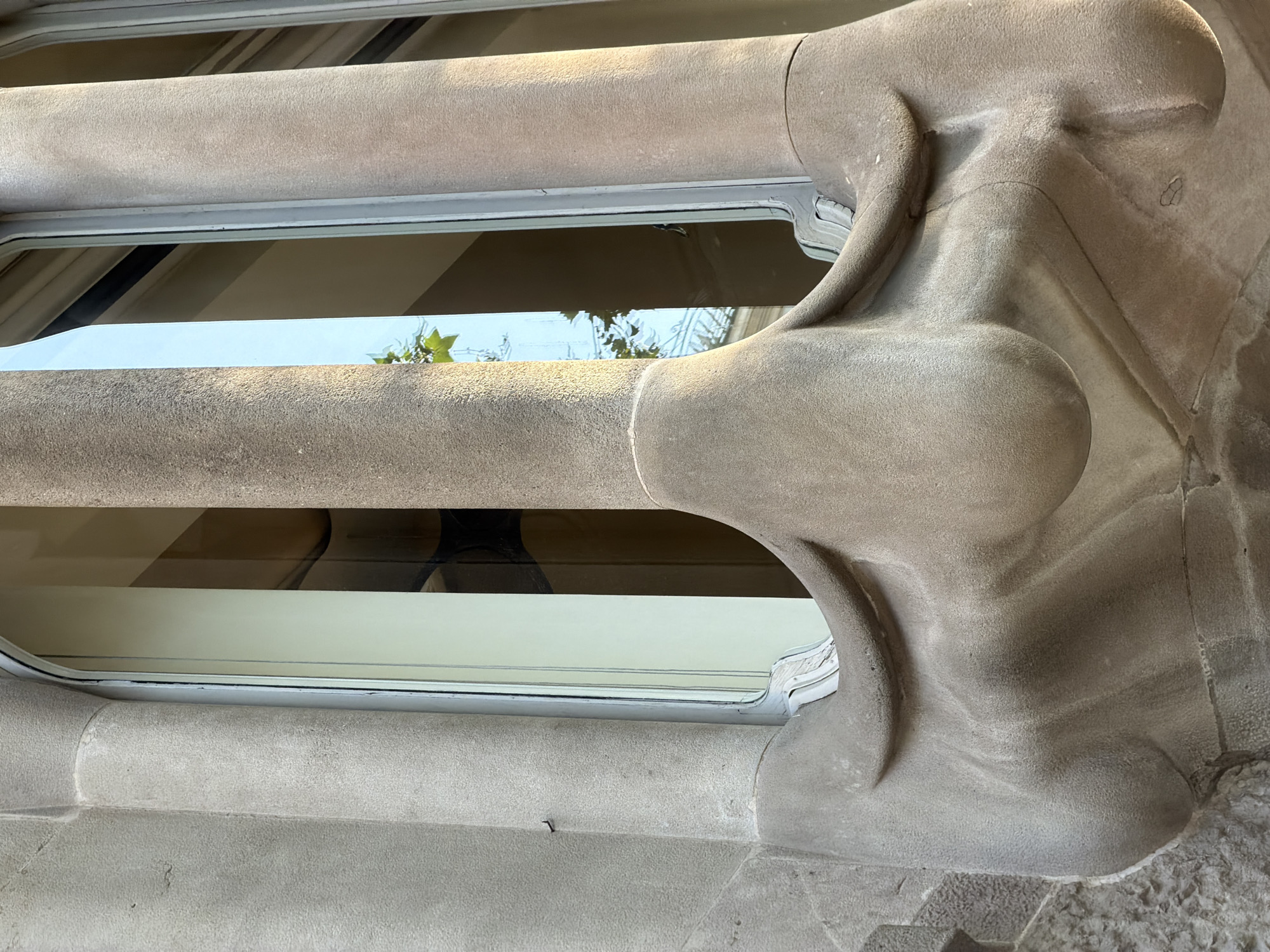 This close-up, oblique photograph captures a distinctive architectural detail from a building in Barcelona, Spain, strongly reminiscent of Antoni Gaudí's work, such as Casa Batlló. The primary subject is a section of a window frame or balcony railing crafted from a light brown, rough-textured stone or concrete. The material is sculpted into organic, flowing, and curvilinear forms that evoke natural elements or bone-like structures.
Two horizontal, elongated elements form the top and bottom of a window opening, separated by a pane of glass. The granular surface of the stone is highly visible, showing subtle undulations and the hand-crafted nature of the design. Through the glass, reflections reveal glimpses of green foliage and branches, indicating an outdoor, natural setting. Faintly visible behind the glass are interior elements, possibly striped blinds or shades, and darker areas.
The scene is illuminated by bright daylight, suggesting a clear day with soft shadows. No people or specific activities are depicted, emphasizing the unique artistry and textural quality of the architecture. The intricate design and naturalistic aesthetic are characteristic of Catalan Modernisme. There is no visible text in the image.
