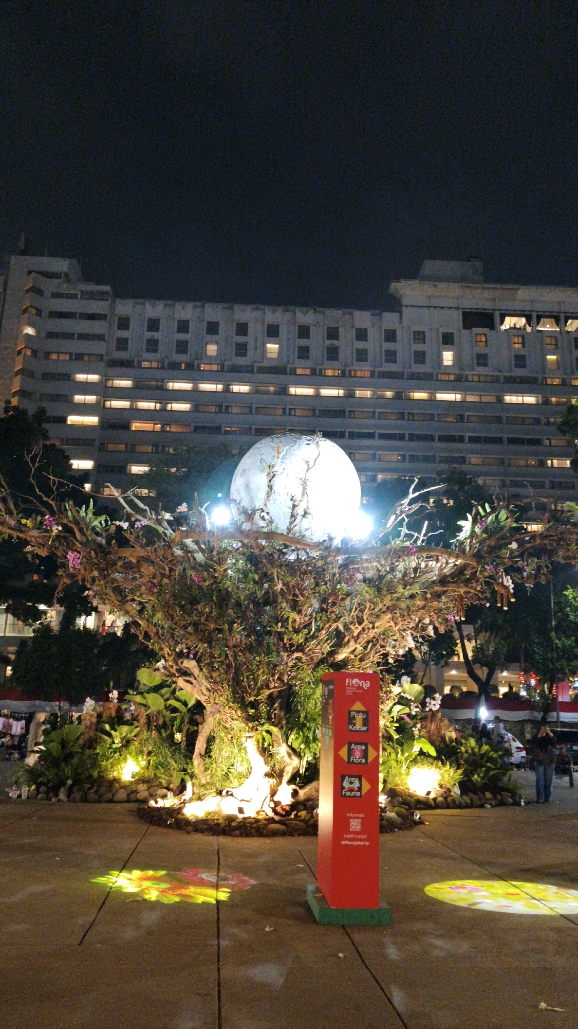 The image shows an outdoor night scene in Central Jakarta, Indonesia, in front of a large, illuminated building. In the foreground, a large, spherical object resembling the moon is suspended above a decorative tree installation with lights and flowers. A red signpost with text and logos is positioned to the right of the tree. The ground in front of the installation is illuminated with colorful projected patterns of flowers. Several people are visible in the background, some standing and some walking. The building behind the installation is a multi-story structure with many windows, some of which are lit. The sky is dark, indicating nighttime.