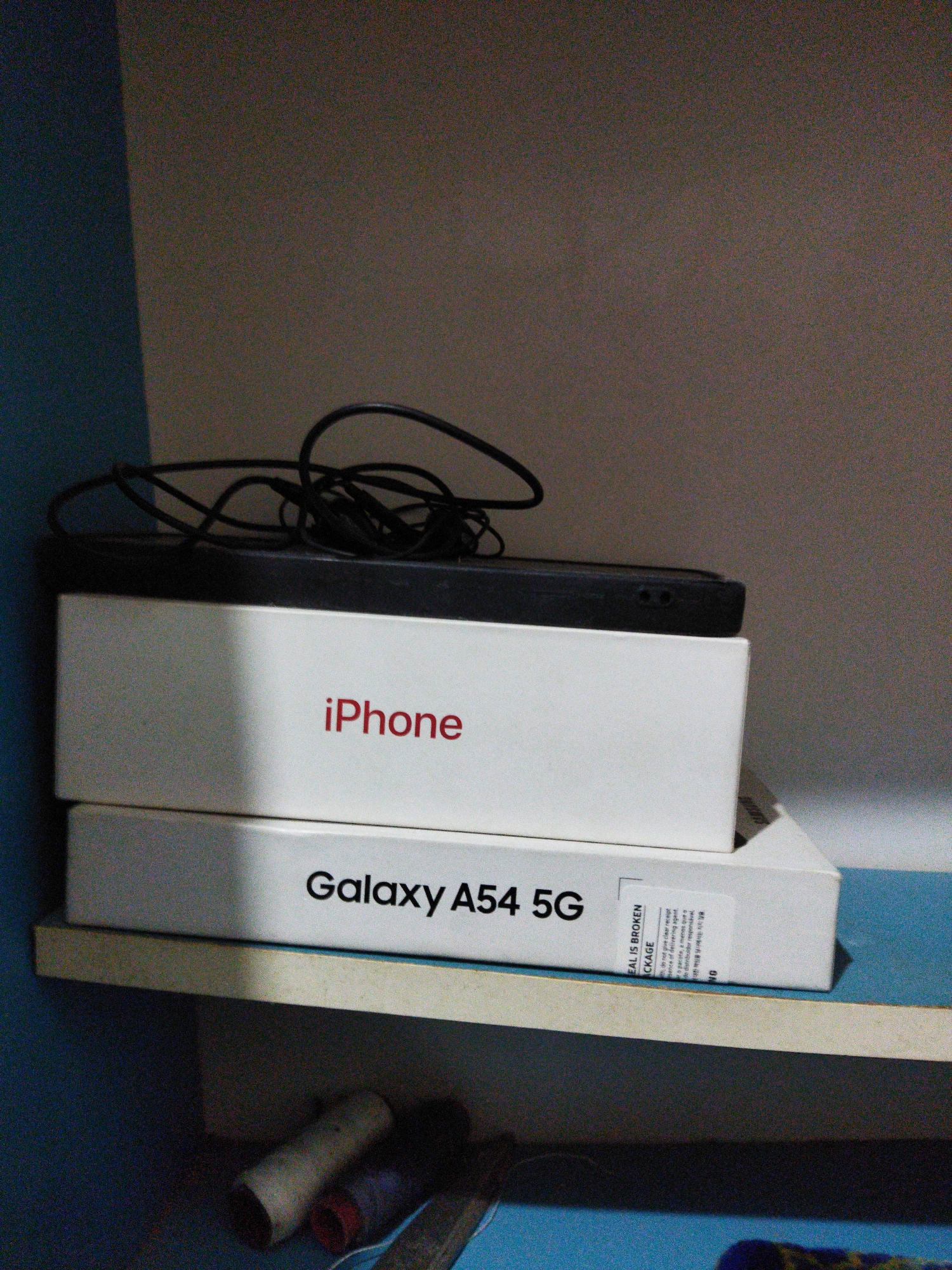 Two white mobile phone boxes are stacked on a shelf. The top box is labeled "iPhone" in red text, and the bottom box is labeled "Galaxy A54 5G" in black text. A black power cord is resting on top of the iPhone box. The shelf is light brown and appears to be made of particleboard. The background is a light gray wall. The image was taken in Bandar Sakti, Indonesia.