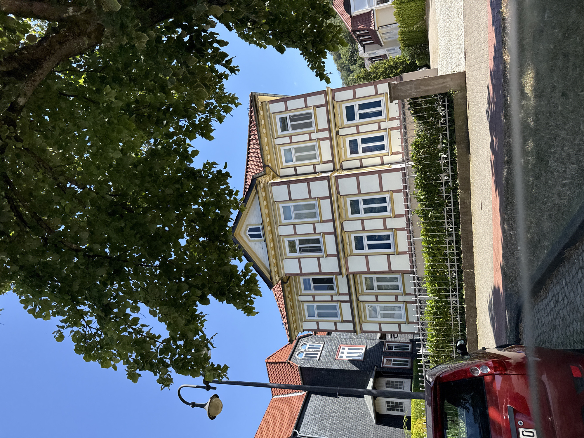 This image depicts a street scene in Bad Lauterberg im Harz, Germany. A red car is partially visible in the foreground on the left, parked on a cobblestone surface. Behind the car and a white metal fence, a traditional German half-timbered house stands out with its cream-colored walls and dark brown beams. The house has a red tiled roof and multiple white-framed windows. To the left of the half-timbered house is another building with a dark gray, scaled facade and a red tiled roof. The sky is clear and blue, and the scene is partially shaded by the branches and leaves of a large tree in the upper portion of the image. A vintage-style lamppost is also visible on the left side of the frame, standing near the buildings. The overall impression is of a well-maintained residential street in a German town.