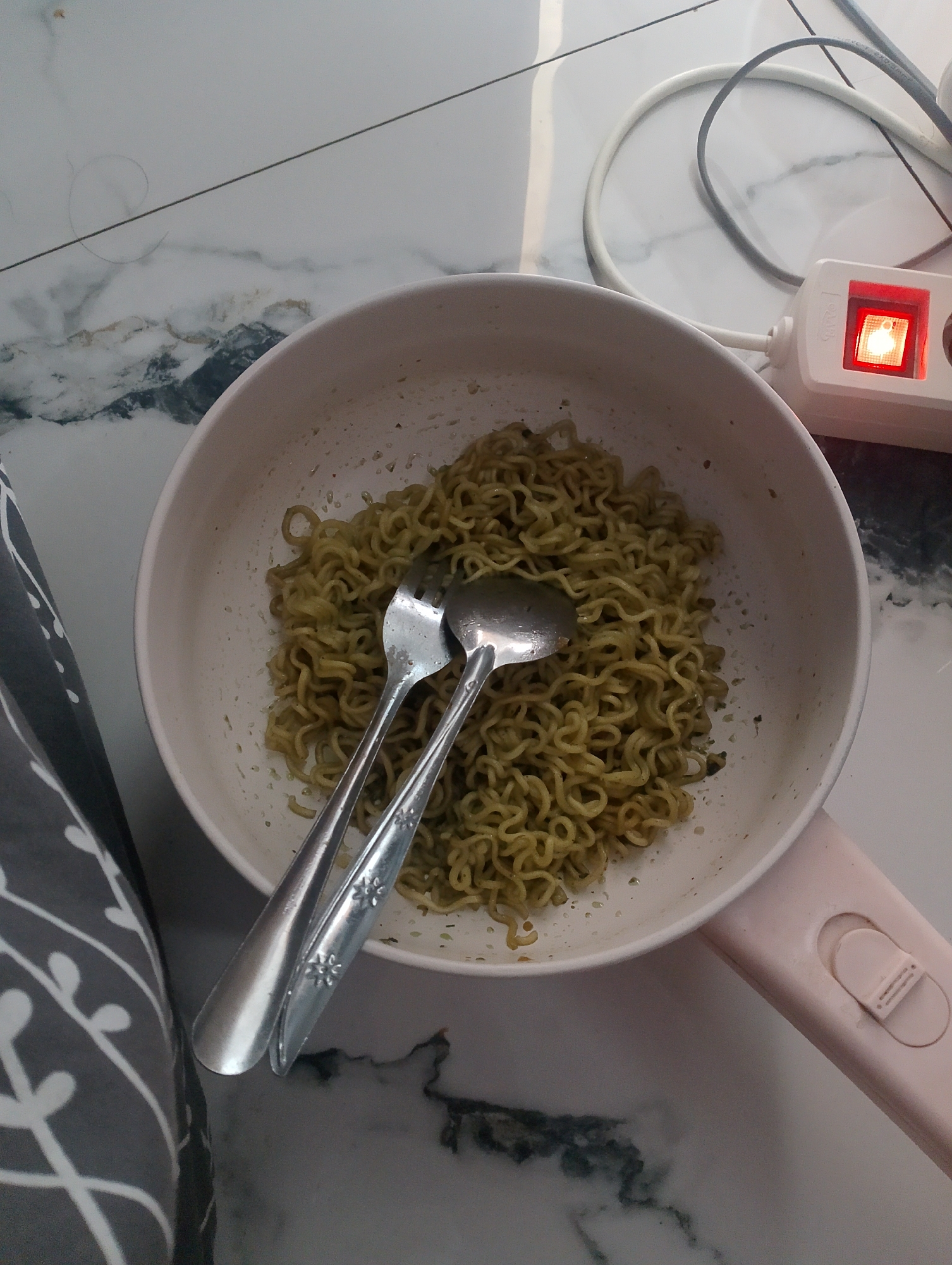 The image shows a close-up, high-angle view of a white ceramic bowl filled with cooked instant noodles. Two metal utensils, a fork and a spoon, are placed in the bowl, on top of the noodles. The bowl is resting on a pink handle, suggesting it is part of a small electric pot or pan. The scene is set on a marble-patterned surface. In the background, a white extension cord with a red illuminated power switch is visible. The environment appears to be indoors, possibly a kitchen or dining area. No people are present in the image. The lighting suggests it is daytime. The location is not specified as Magelang, Indonesia.