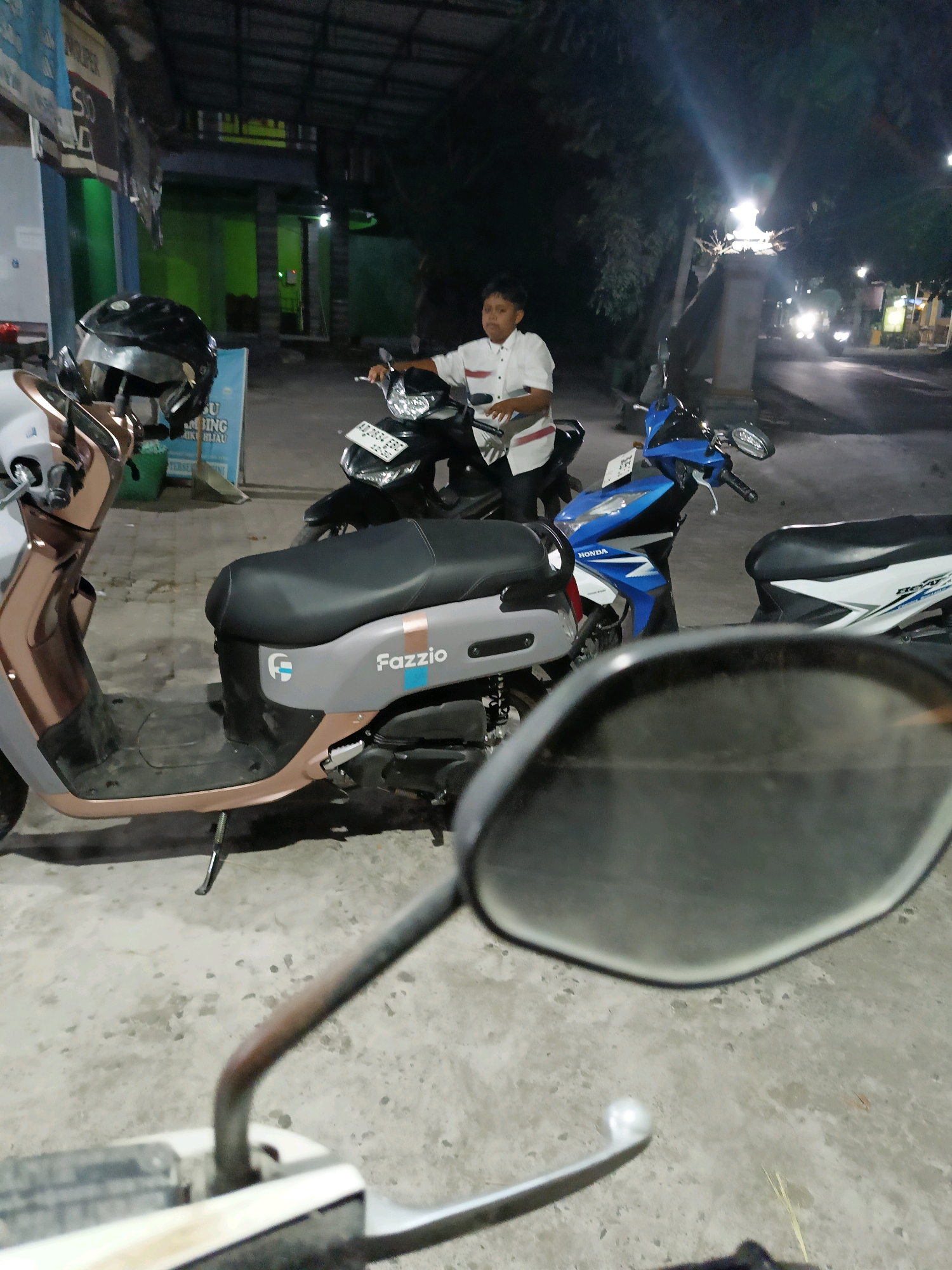 The image depicts a nighttime scene on a street in Manisrenggo, Indonesia, likely a residential or commercial area. In the foreground, a person is partially visible in the reflection of a motorcycle's rearview mirror. A grey and copper colored scooter with the "Fazzio" logo is prominently parked on the left. Behind it, a young boy wearing a white shirt with red stripes is standing next to a black motorcycle. Further back, a blue Honda motorcycle is parked, and to its right, a white and black motorcycle is visible. The background shows buildings with illuminated windows and streetlights, and trees lining the street, suggesting an urban or semi-urban setting at night. The ground is paved with concrete.