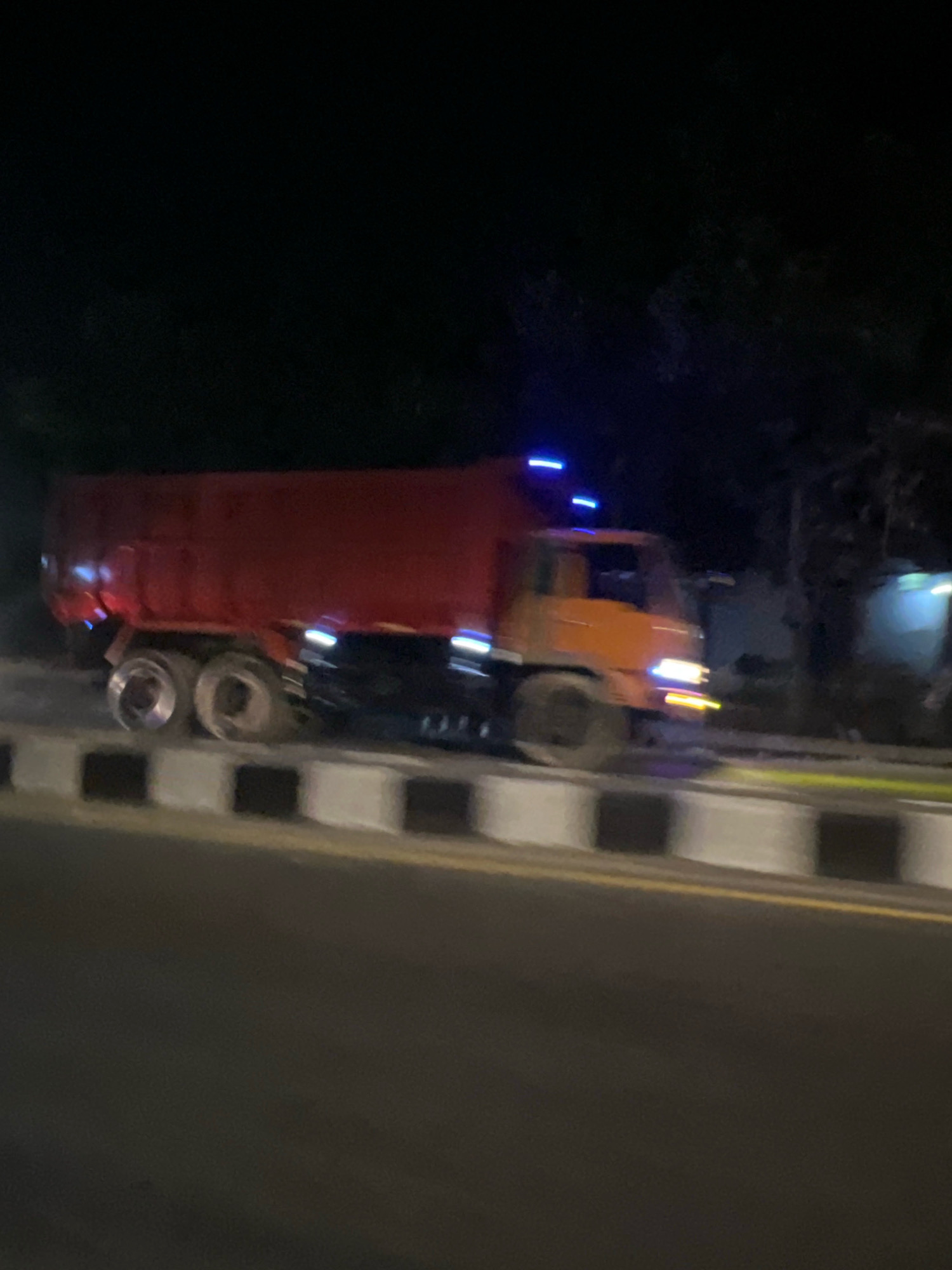 A red dump truck with an orange cab is driving on a road at night. The truck has multiple wheels and blue lights on its roof. A concrete barrier with alternating black and white squares lines the side of the road. Trees and a building are visible in the background. The image appears to be taken in Salam, Indonesia.