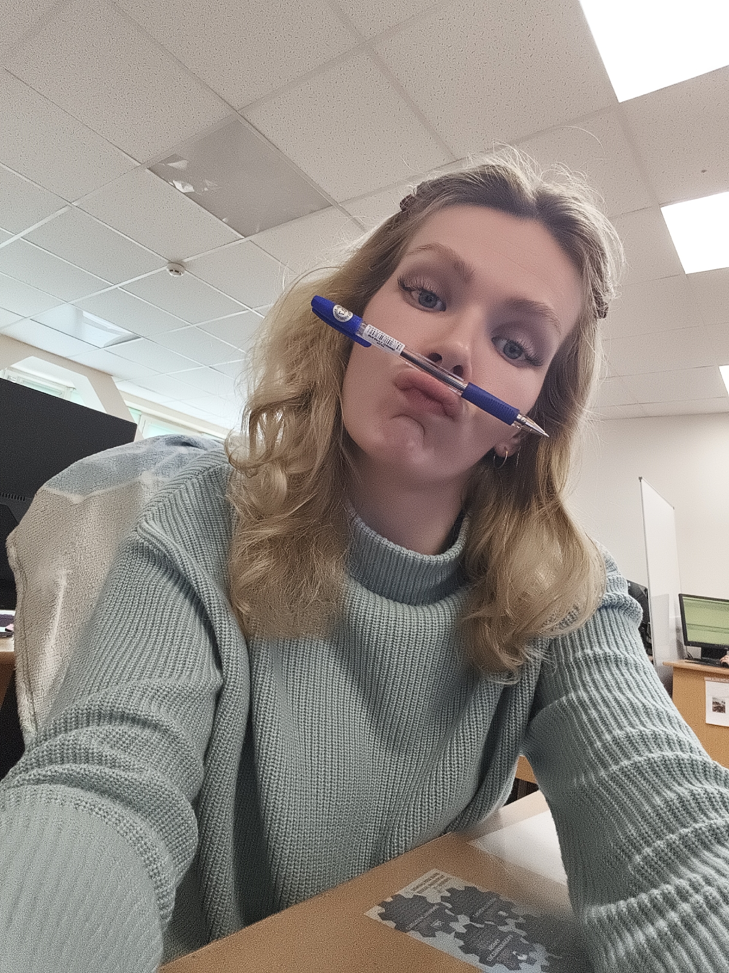 A young woman is positioned in an office setting, evident from the fluorescent ceiling lights and office furniture visible in the background. She is holding a blue ballpoint pen horizontally between her nose and upper lip, her expression playful with slightly pursed lips. She is wearing a light blue, ribbed knit sweater with a turtleneck. Her blonde hair is styled with a few strands pulled back, and she is wearing small hoop earrings. In the background, there is a computer monitor and a divider screen, typical of an office environment. There is also a desk with an item that seems to have a sticker with a logo on it. The location in the city of Unknown, Unknown is impossible to verify from the given image.