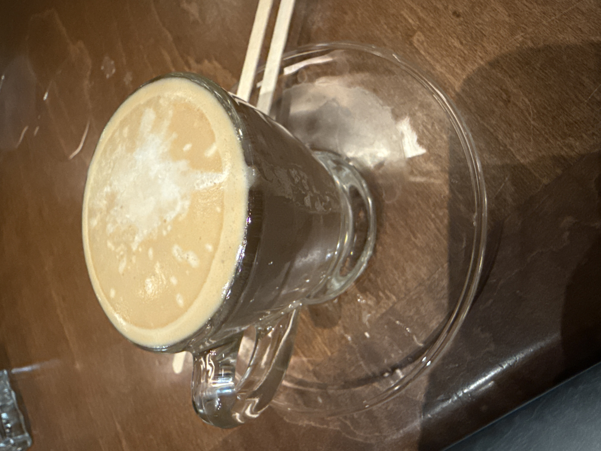 A close-up overhead shot shows a glass mug of coffee sitting on a clear glass saucer atop a wooden table. Two tan-colored stir sticks are partially visible behind the mug. The coffee has a light tan foamy top. The mug handle is clear glass. Light reflects off the glass and the polished table surface. The photo was taken during the day. It is possible this image was taken in Dania Beach, United States of America.