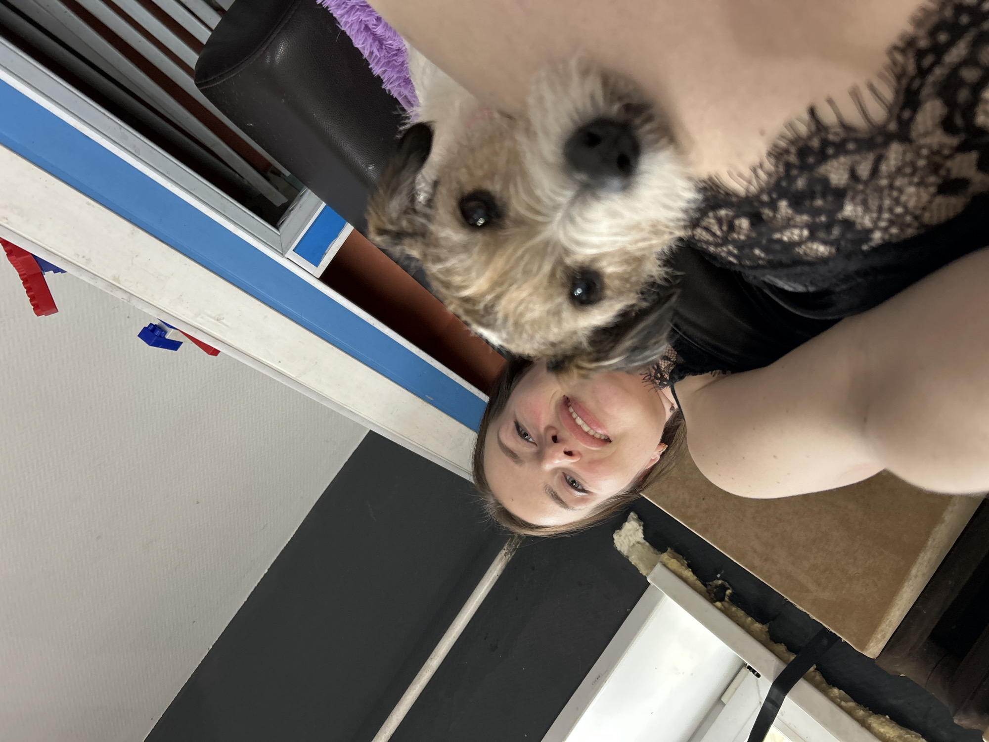 The image depicts a woman holding a small terrier-type dog close to her face. The woman is smiling and looking at the camera. She is wearing a black top with lace detailing. The dog is positioned between her shoulder and face. In the background, elements of a room are visible, including a window frame with blue and white trim, a black and white wall, and part of a black chair with purple fabric. The location is not discernible as Rusalevka, Russia without further information or visual cues.
