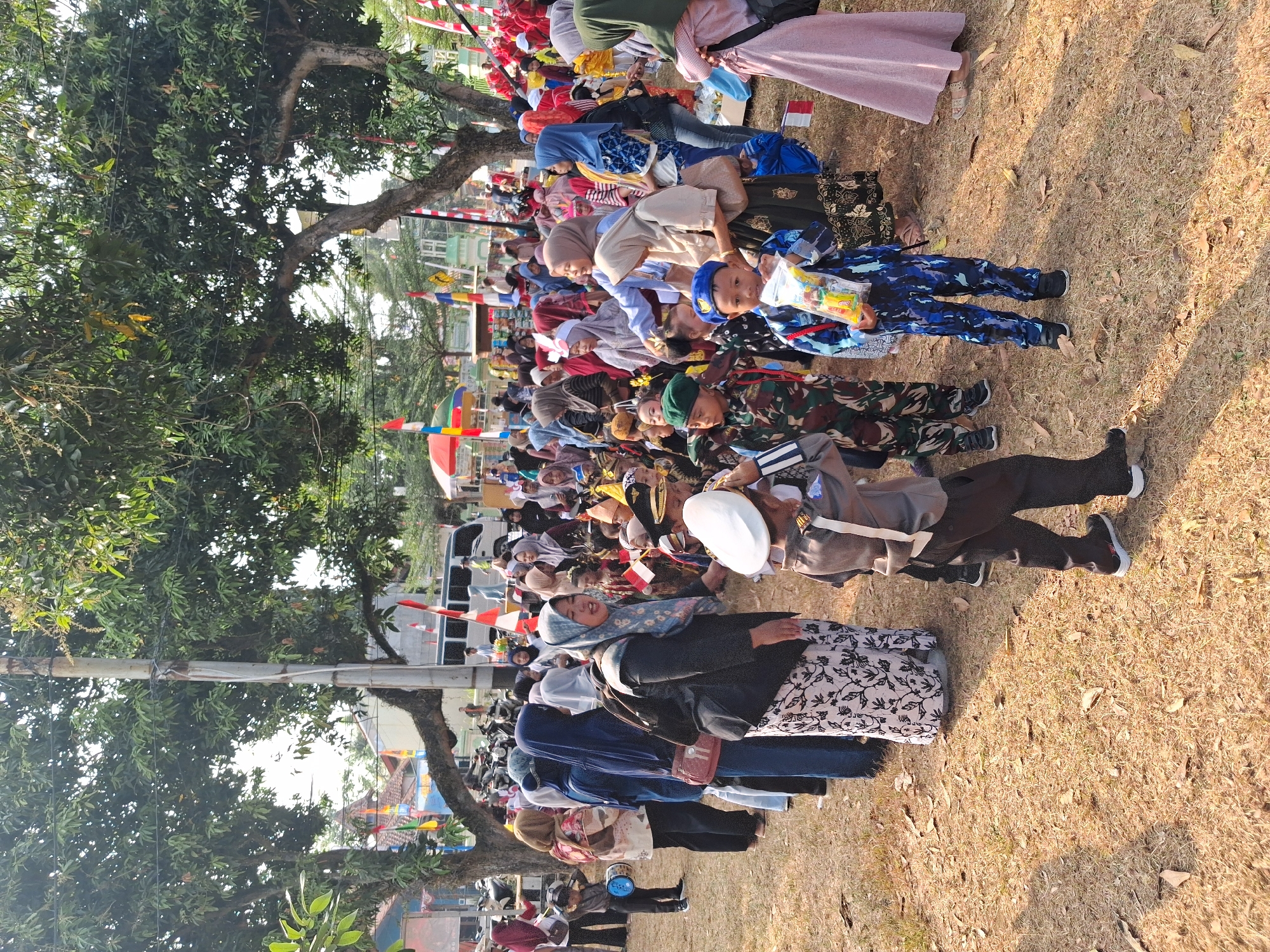 The image depicts a gathering of people, primarily children dressed in various costumes, in an outdoor setting in Balapulang, Indonesia. The ground is covered with dry grass and fallen leaves, suggesting a warm, dry climate. Several large trees with dense foliage provide shade, and a utility pole is visible on the left side of the frame. In the background, a white van and a row of motorcycles are parked.
Many of the children are wearing what appear to be military or ceremonial uniforms, including camouflage fatigues, police outfits, and some with hats and berets. One child in the foreground is wearing a blue patterned uniform and holding a small bag of candy. Another child, dressed in a police uniform, is saluting. Adults are also present, some wearing hijabs and traditional Indonesian attire. Small Indonesian flags are visible throughout the scene, indicating a national celebration or event. The overall atmosphere appears to be festive, with people mingling and children participating in some form of activity or parade.