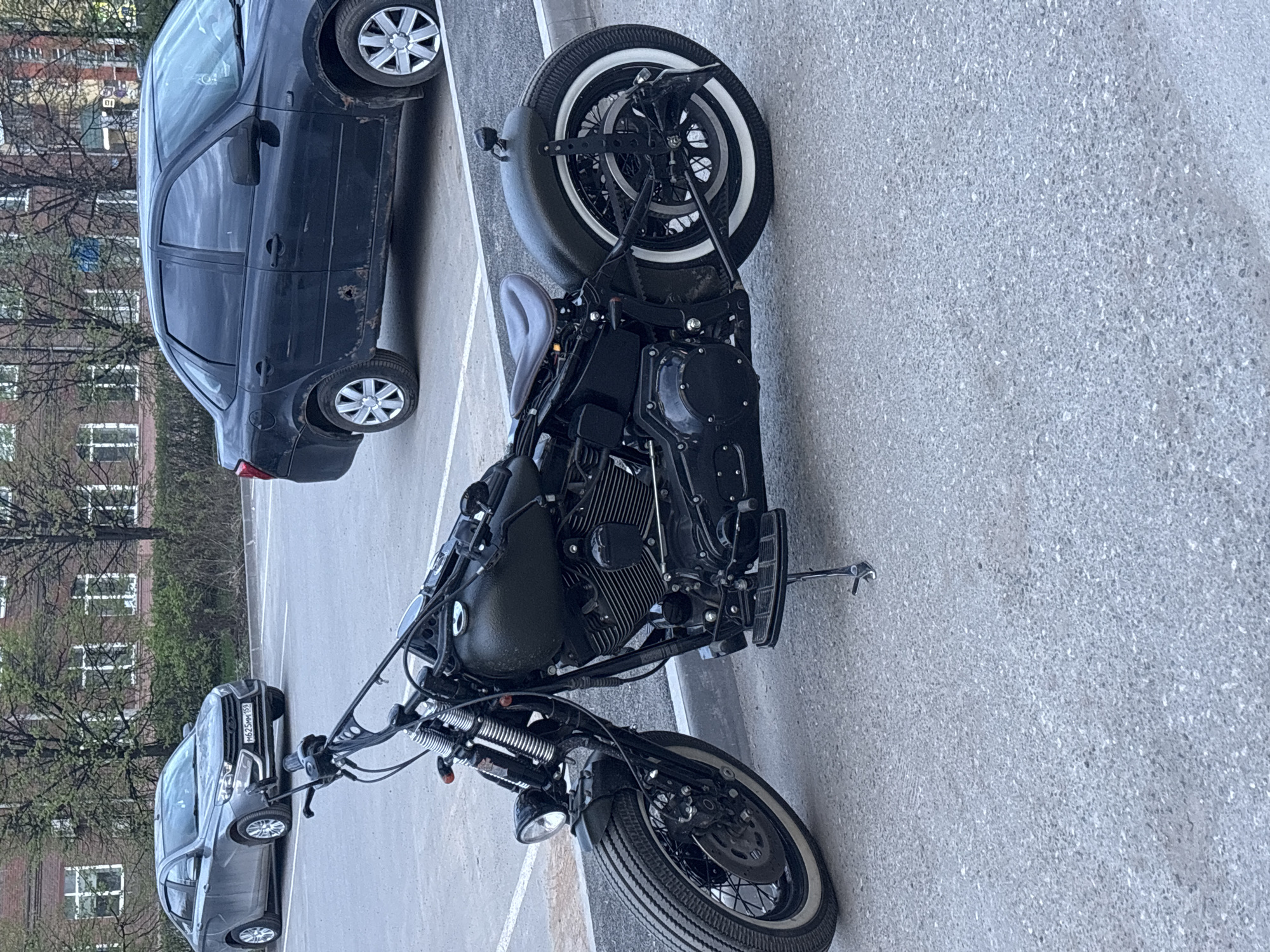 The image depicts a black motorcycle parked in a parking lot in Perm, Russia. A black sedan is parked alongside the motorcycle on the left of the frame, and another car is further away from the camera. The motorcycle has a white-walled front tire and a side-mounted license plate. The parking lot is paved with a gray surface. Buildings with trees are in the background, suggesting an urban environment.