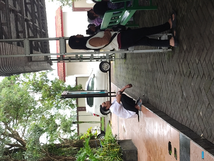 This image captures an outdoor or semi-outdoor waiting area, likely during daytime under overcast conditions, given the soft lighting and lack of strong shadows. The ground is primarily paved with a herringbone brick pattern, appearing somewhat damp, possibly from recent rain or cleaning.

In the foreground, a person wearing a white shirt, dark pants, and flip-flops is seated on the ground next to a low curb or planter box, looking down or towards the right. Behind them, under a metal shelter structure, a woman stands upright, facing to the right. She has long dark hair and is dressed in a light-colored top (beige and white) and black pants, also wearing flip-flops. Further back, obscured by the standing woman, other individuals are visible sitting on a green bench, indicating a waiting area.

In the midground, a white car is parked, and a dark-framed display stand is positioned near it. The background reveals part of a building with a reddish-brown roof and white walls, partially hidden by abundant green trees and foliage, especially on the left side of the frame. The overall atmosphere suggests a quiet, patient wait in a public or institutional setting. No legible text is visible in the image.