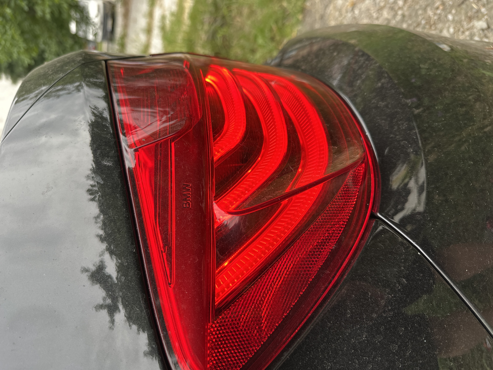 The image shows a close-up, rear-side view of a dark-colored BMW car, focusing on the taillight. The taillight is illuminated with a bright red LED pattern. The car's bodywork appears dusty. In the background, out of focus, is greenery and a sliver of a white vehicle parked on a dirt or gravel surface. The setting suggests an outdoor environment, possibly a residential area or parking lot, in Unknown, Unknown.