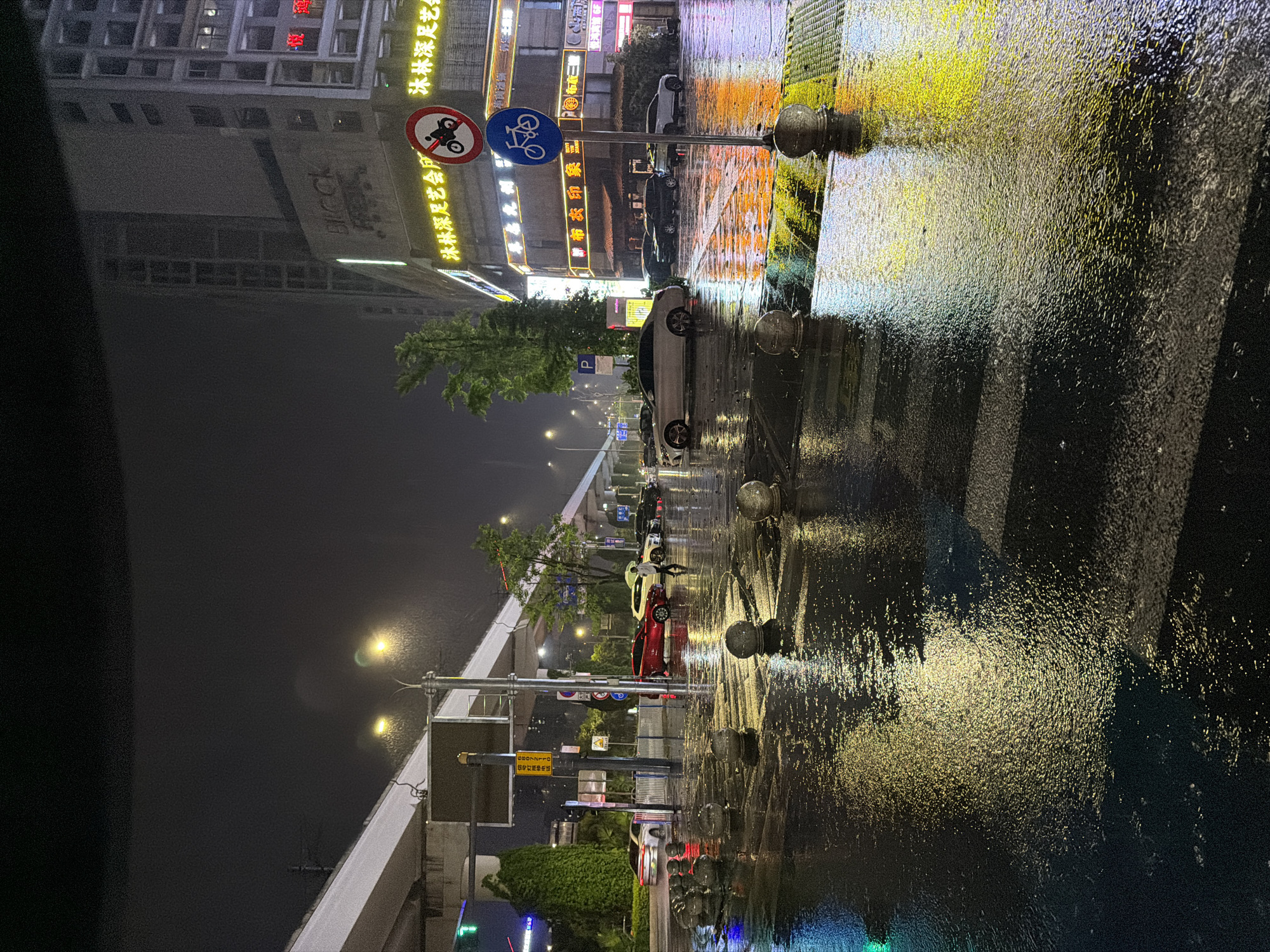 The image captures a rain-soaked street scene in Chongqing High-tech Zone, China, at night. The street is illuminated by streetlights and neon signs, which reflect off the wet pavement. The ground is covered in a layer of water, creating a shimmering effect that obscures the details of the road surface. The image shows parked cars and signs indicating no motorcycles allowed. In the distance, a building with the word "BLOCK" is visible. Street signs in Chinese are shown in the scene.