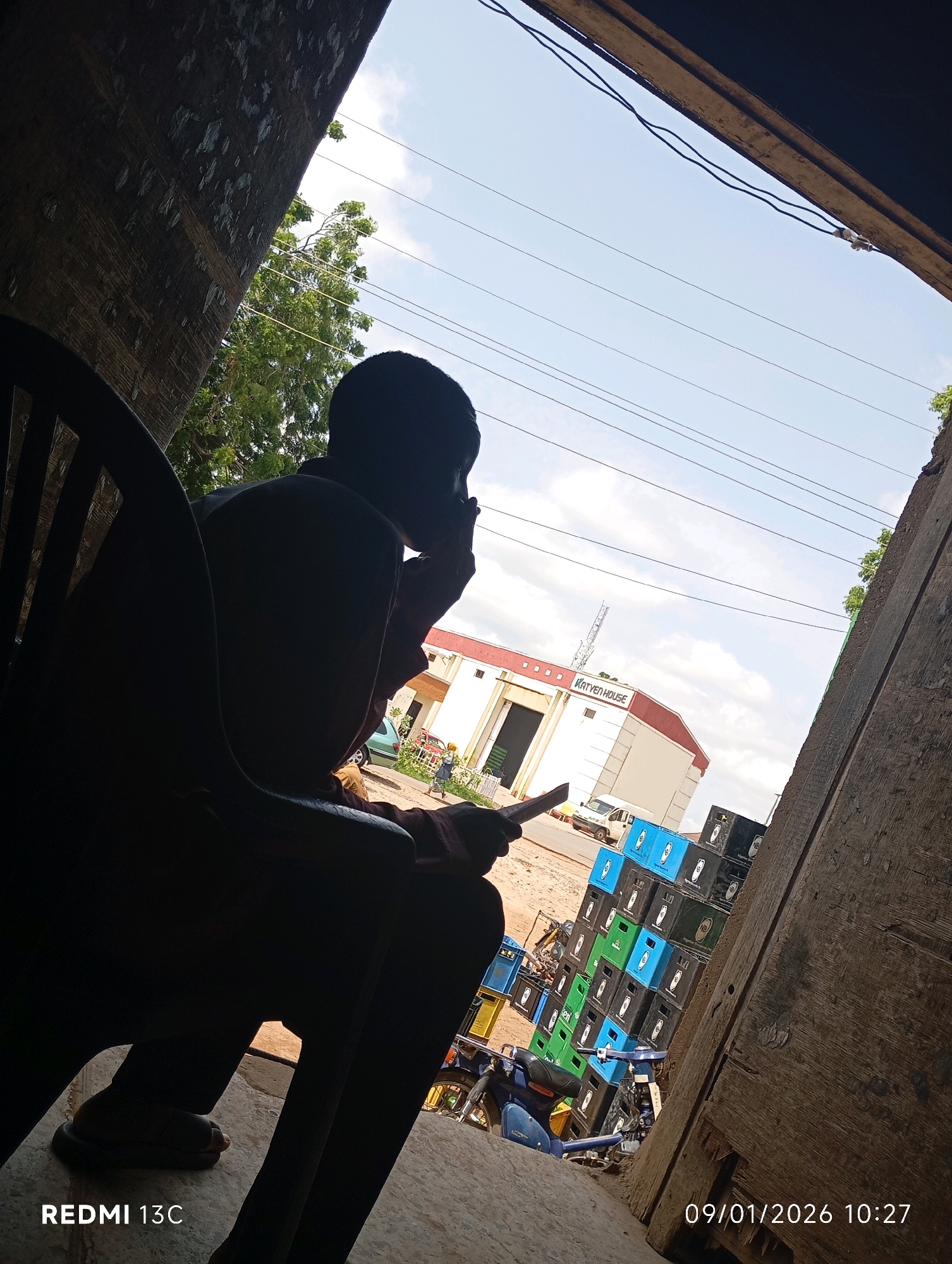 The image depicts a scene in Shendam, Nigeria, captured on January 9, 2026, at 10:27 AM. In the foreground, a person is seated in a dark chair, silhouetted against the bright sky. The individual is holding a mobile phone or similar device and appears to be engaged with it. Their legs are visible, wearing sandals.
The background reveals a commercial building with red and white signage that reads "KATYEN HOUSE." Outside the building, stacks of colorful crates, likely for beverages, are piled high. A white van and a blue motorcycle are also present, suggesting commercial activity or transportation. The environment appears to be an outdoor space, possibly a market or a commercial hub, with visible electrical wires overhead and a cloudy sky. The perspective is from within a covered area, looking out towards the street.