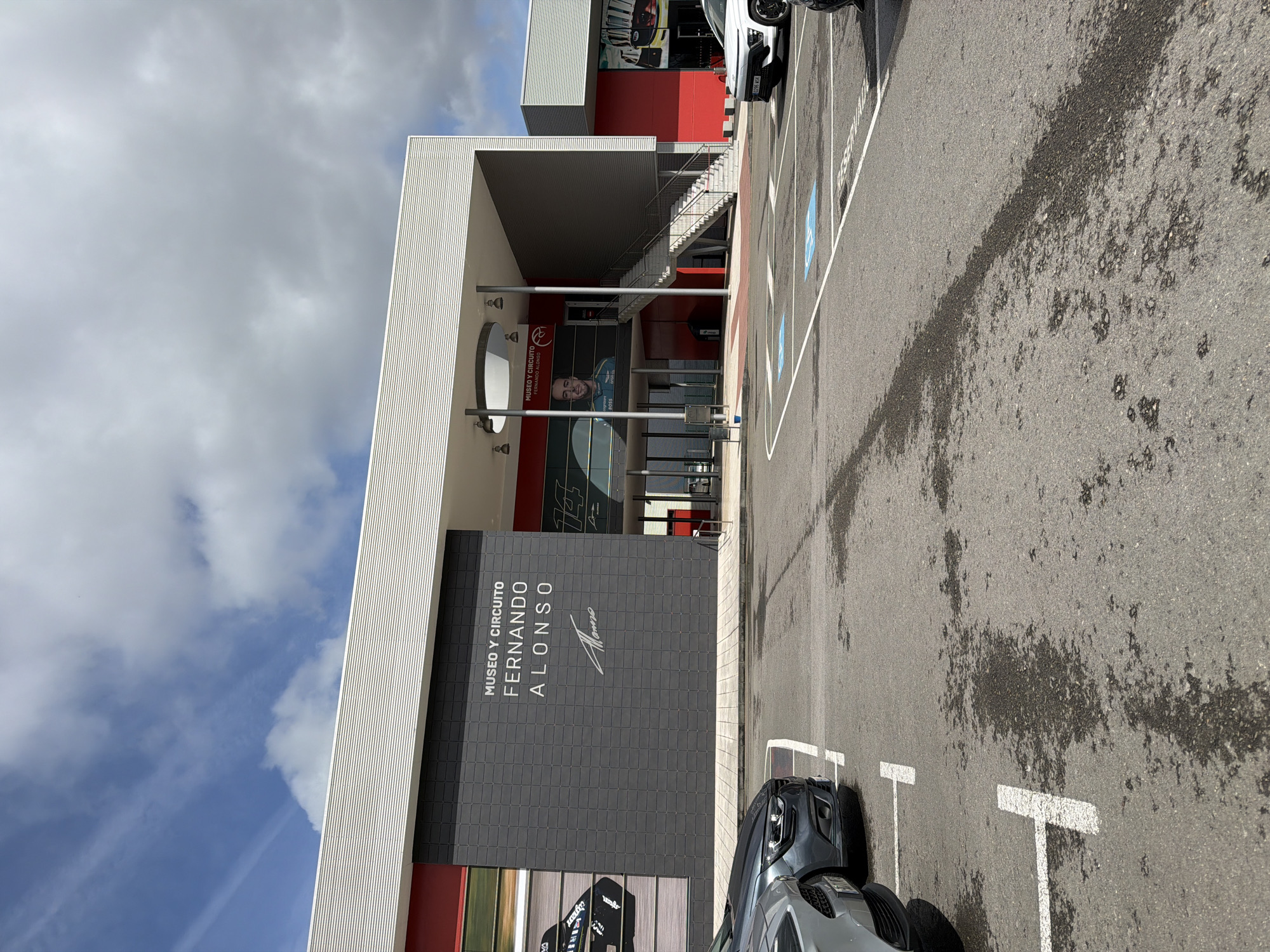 The image depicts the exterior of the Museo y Circuito Fernando Alonso, located in Llanera, Spain. The modern, multi-story building features a facade with alternating sections of white corrugated metal, red panels, and dark gray square-tiled surfaces. The museum's name, "MUSEO Y CIRCUITO FERNANDO ALONSO," is prominently displayed in white lettering on a dark gray section of the building, accompanied by Fernando Alonso's signature. The main entrance, centrally located, is recessed and features glass doors, above which a large banner displays a portrait of Fernando Alonso in racing attire. An external staircase with a metal railing ascends to an upper level on the right side of the main entrance. Large graphic images of racing cars and helmets are visible on other parts of the building's facade.
In the foreground, an asphalt parking lot is visible, showing white painted lines delineating parking spaces. Several wet patches or stains are present on the asphalt surface. A silver/gray car is partially visible in the immediate left foreground, followed by a dark gray car (a Renault Mégane) parked to its right. A white Hyundai Kona is parked further right, with another white car partially visible on the far right. Two parking spaces are marked with blue wheelchair accessibility symbols. The sky above is partially cloudy, featuring patches of blue sky and scattered white to light gray clouds, with a faint contrail visible in the upper left. No individuals are visible in the scene.