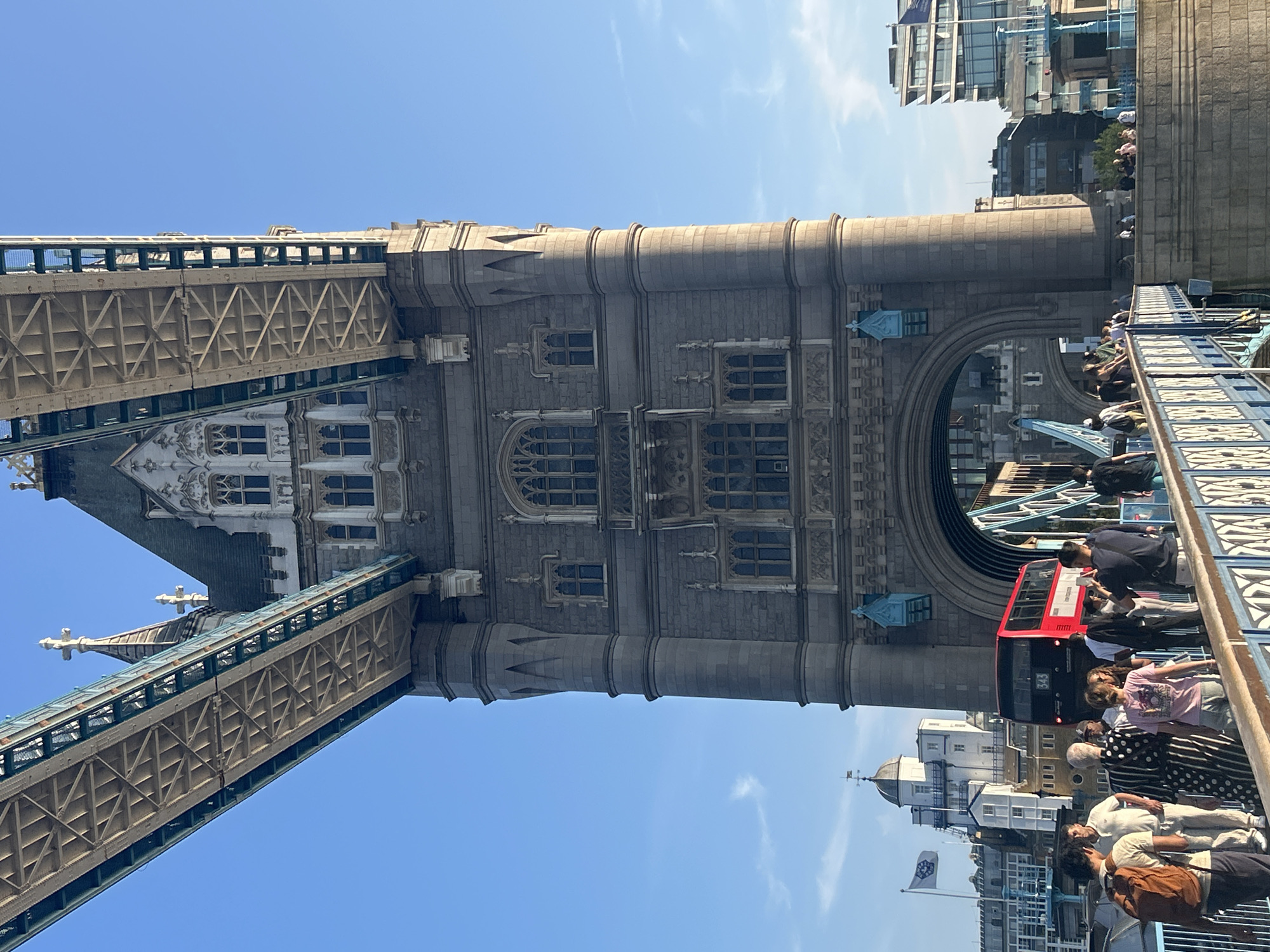The image depicts a low-angle, full-frame shot of the Tower Bridge in London, United Kingdom, on a clear, sunny day. The iconic structure, with its grey stone towers and intricate metalwork, dominates the scene. A red double-decker bus, numbered 343, is visible passing through one of the bridge's arches. Several people are walking along the pedestrian walkway of the bridge, some of whom are looking at their phones or taking pictures. The sky is a bright blue with a few wispy clouds. In the background, other buildings and structures are visible, including a white building with a domed roof and a modern glass-fronted building. The overall atmosphere is one of a bustling tourist attraction on a pleasant day.