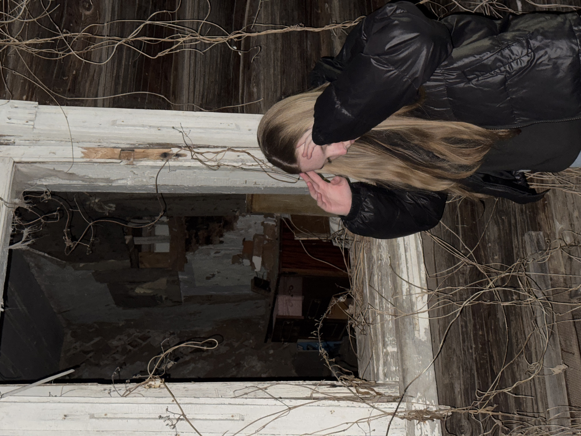 The image shows a young woman looking into an abandoned building. The building appears to be in a state of disrepair, with damaged walls and decaying wood. The woman is wearing a black puffer jacket and covering her eyes with her hands. Dry vines are growing around the window frame. The location of the building is Zhigalki, Russia.