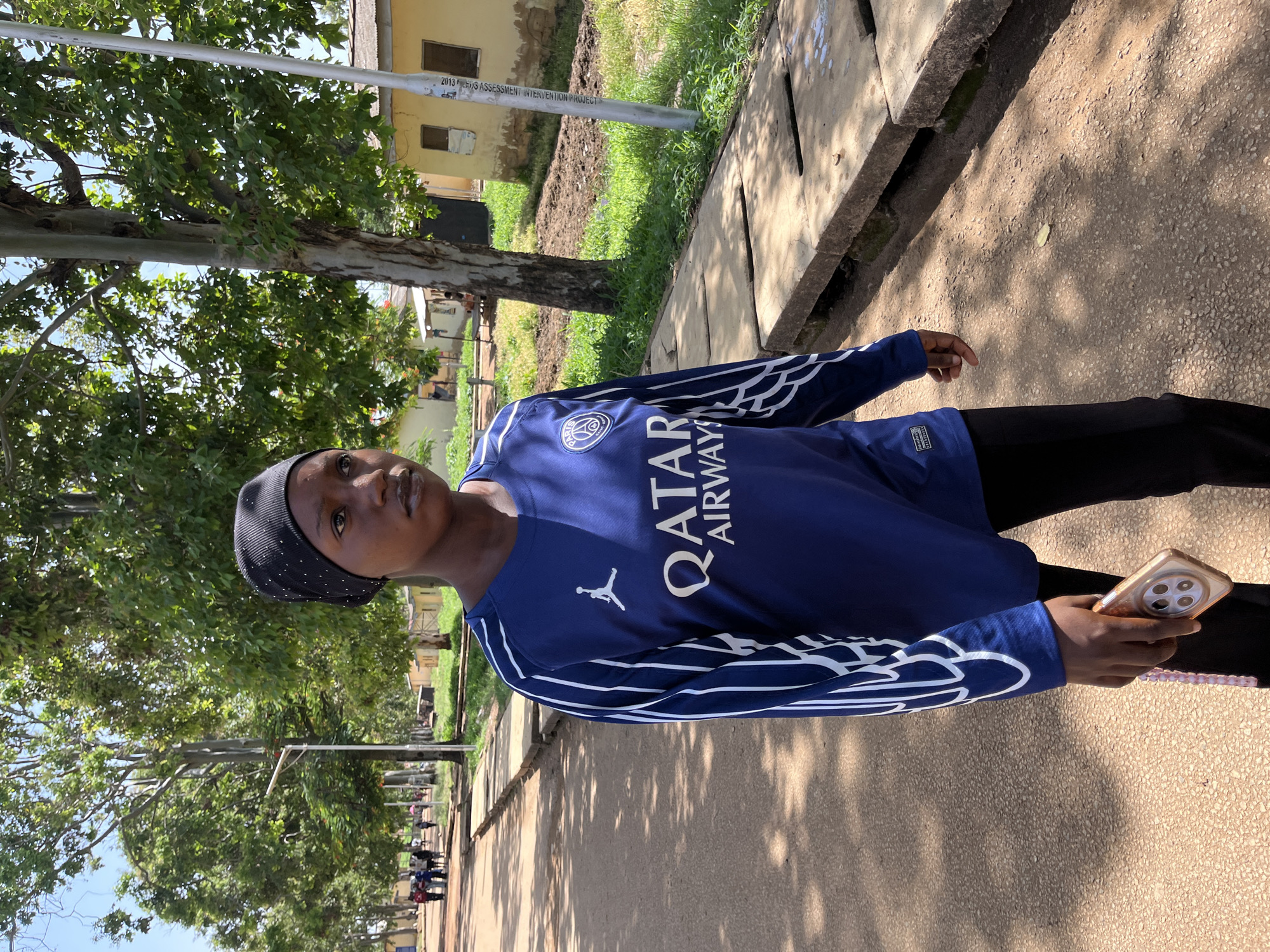 An outdoor, full-body portrait captures a young person standing on a paved path in Rafin Par, Nigeria. The subject, likely female, wears a dark blue long-sleeved shirt featuring the Paris Saint-Germain (PSG) crest, a white Jordan logo, and the sponsor text "QATAR AIRWAYS". She also wears black pants and a black head covering with small white polka dots. Her right hand holds a light-colored smartphone with multiple camera lenses and a pink beaded strap attached.
The background features lush green trees with dappled sunlight creating shadows on the path. To the right, a concrete drainage ditch runs alongside the path. In the mid-ground, a light yellow building is partially visible, and a metallic utility pole stands upright. Text on the pole in one of the crops reads "2013" and "S ASSESSMENT INTERVENTION PROJECT". Further down the path, in the distance, a few other people can be seen. The bright sunlight and strong shadows suggest it is daytime, likely mid-morning or early afternoon. The overall scene depicts a casual moment in a sunny, tree-lined environment.