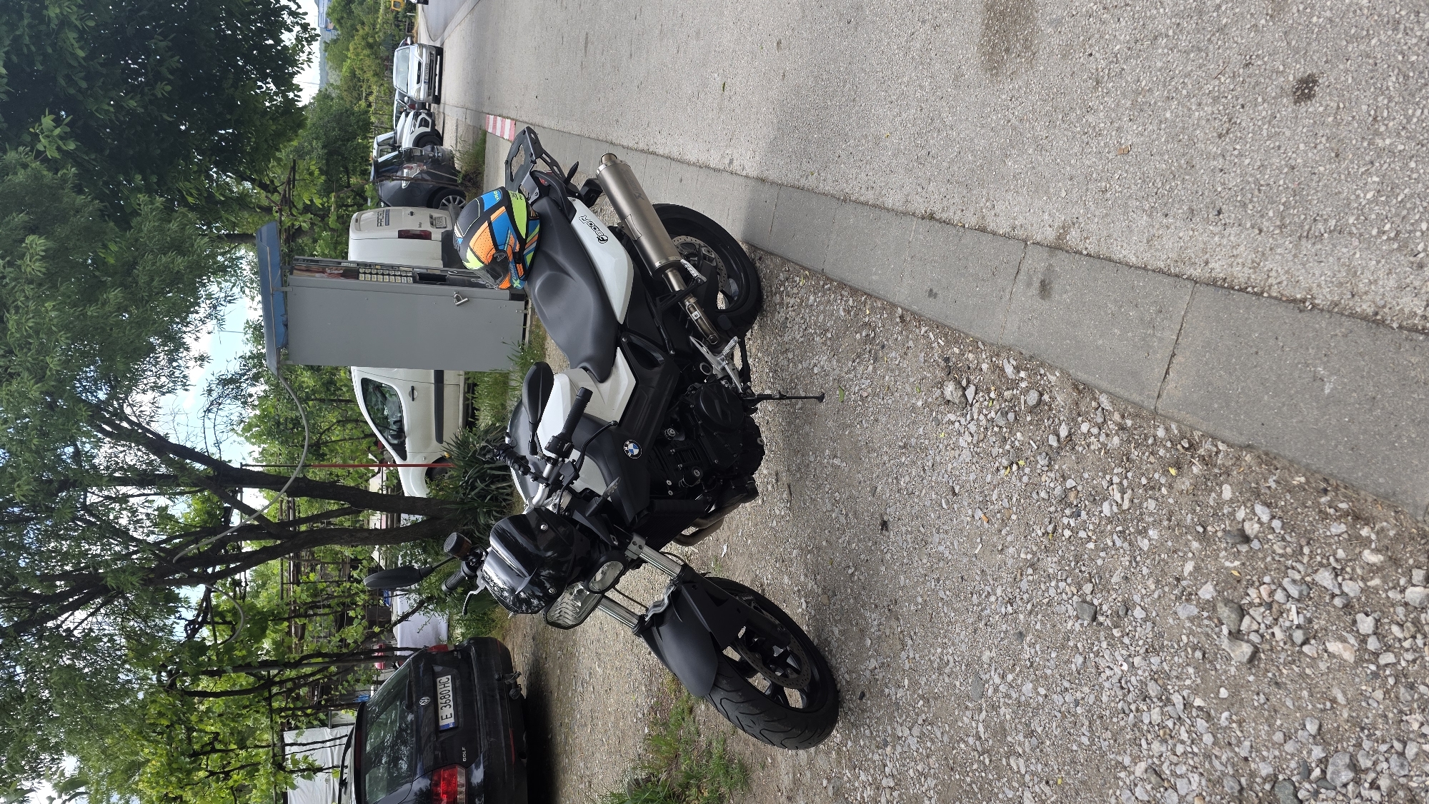 The image shows a parked BMW F800R motorcycle with a helmet on its seat, located on a gravelly patch adjacent to a sidewalk and asphalt road. The location is likely in an urban area, indicated by the presence of other parked vehicles, trees, a utility box, and a black Volkswagen Golf with license plate E 3480 HC visible in the background. The scene is set in Blagoevgrad, Bulgaria based on the vehicle registration, the photograph being rotated at a 90-degree angle. There are no people present in the shot. The image is taken during the daytime with reasonable lighting.