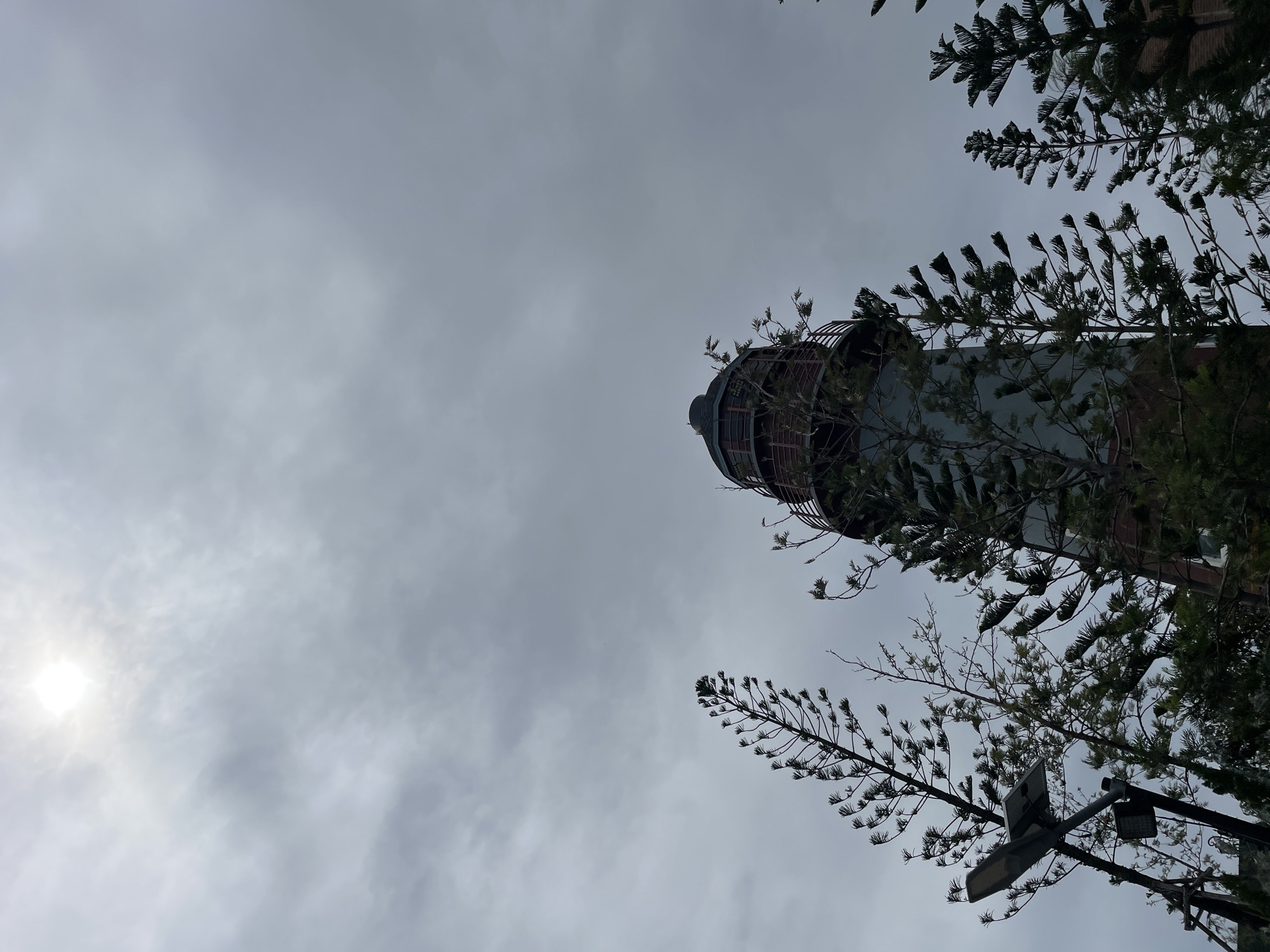 A lighthouse stands tall amidst a cloudy sky in Pattaya City, Thailand. The lighthouse has a cylindrical base with a weathered, rustic appearance, topped by a darker, octagonal structure with windows and a railing. Surrounding the lighthouse are several pine trees with distinctive, feathery needles, some of which obscure parts of the structure. In the foreground, a solar-powered streetlamp is visible on a metal pole, its dark rectangular panel angled towards the sky. The sun is a bright, hazy orb peeking through the thick, gray clouds, casting a diffused light over the scene. The overall impression is one of a somewhat overcast day, with the stark lines of the lighthouse contrasted by the organic shapes of the trees.