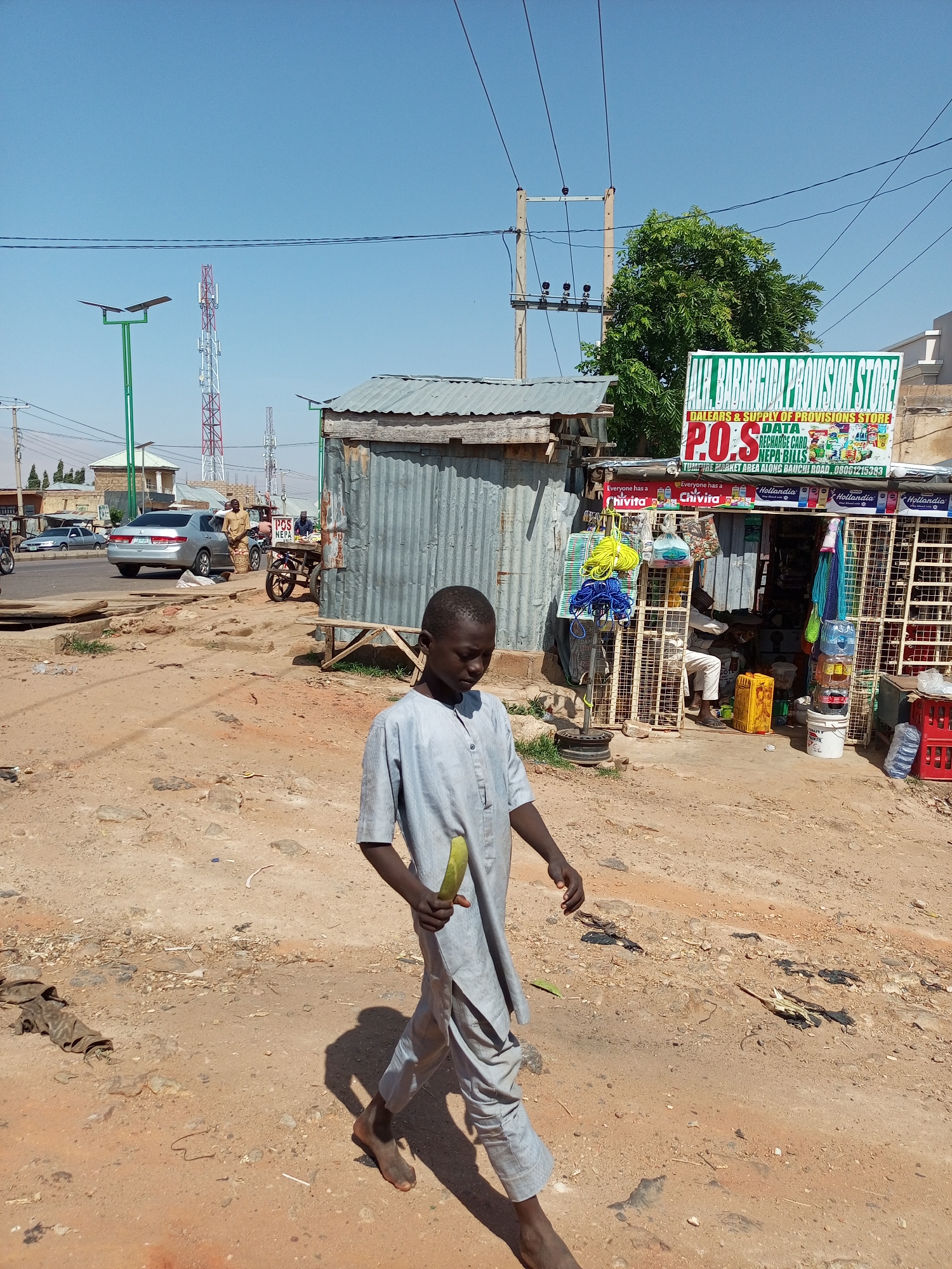 This image depicts a street scene in Gombe, Nigeria, under a clear blue sky, suggesting it is a sunny day. The foreground is dominated by a young boy walking towards the right of the frame, holding a green object that appears to be a vegetable or fruit. He is dressed in traditional light blue clothing.
Behind him, a small provision store, identified by signage as "ALL BABANGIDA PROVISION STORE," is visible. The store has a corrugated metal exterior and displays various products, including drinks, snacks, and other household items. Signs advertise "DATA," "RECHARGE CARD," and "NEPA BILLS," indicating the services offered. The store's location is noted as "TUMFURE MARKET AREA ALONG BAUCHI ROAD."
Further in the background, a paved road is seen with a silver car and a motorcycle. Several communication towers are also present in the distance, along with what appears to be a solar-powered street light. The ground is dusty and unpaved, characteristic of many street environments in the region. The overall scene conveys a sense of daily life and commerce in a bustling Nigerian market area.