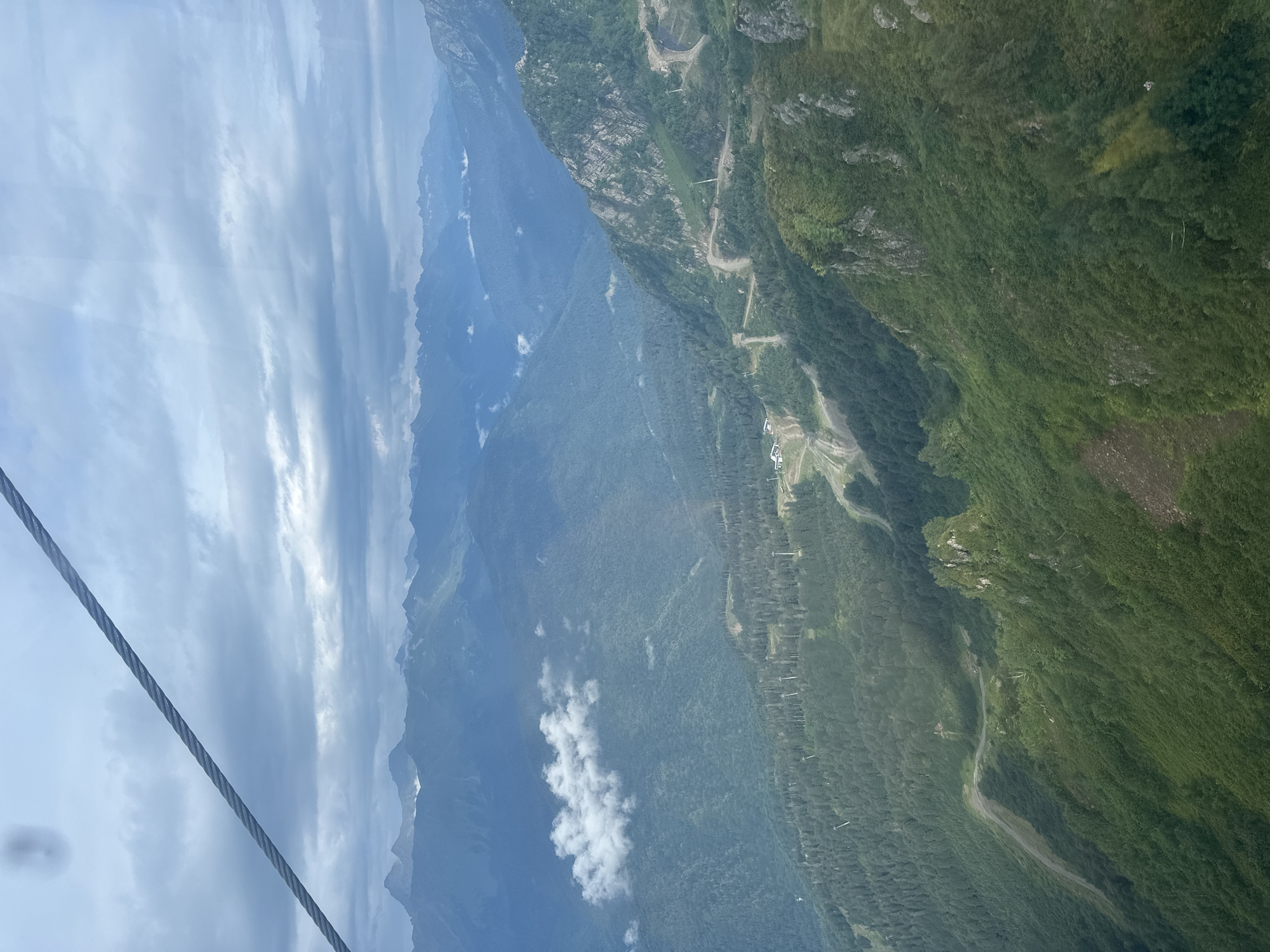 The image shows a panoramic view of a mountainous landscape, likely taken from a cable car descending towards a ski resort area. In the foreground, a thick steel cable, part of the cable car system, stretches across the upper left quadrant of the frame. Below the cable, the terrain is characterized by steep, densely forested slopes and dramatic mountain peaks receding into the distance. The vegetation is lush green, indicative of a warm season.
Several roads and paths are visible winding through the mountainside, some appearing to be ski trails or service roads for the resort. Small buildings, likely associated with the ski resort infrastructure, are scattered throughout the middle ground. Clouds are present in the sky, with some patches of white clouds floating amongst the distant mountain ranges, casting shadows on the slopes below. The overall lighting suggests an overcast or partly cloudy day, with diffused sunlight.
The location is identified as Estosadok, Russia, a resort town in the Krasnodar Krai region known for its ski resorts in the Caucasus Mountains. The scene depicts a typical view from one of the cable car routes in the area, offering a sweeping vista of the natural beauty and the man-made ski infrastructure integrated into the mountain environment. There are no people or animals clearly visible in the image. The primary subjects are the natural mountainous environment, the cable car component, and the resort facilities.