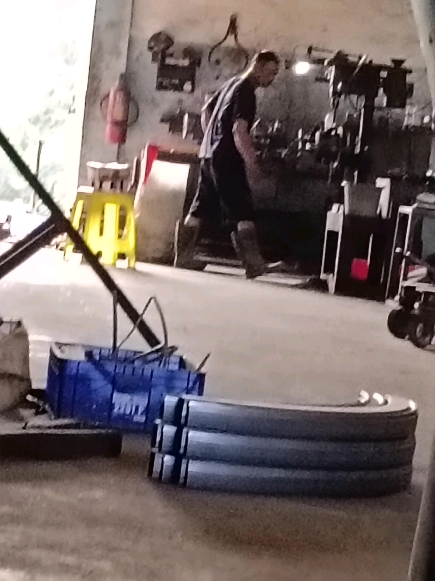 In Belimbing, Indonesia, a man is working in what appears to be a workshop. He is wearing dark clothing and boots and is standing next to a piece of machinery. In the foreground, there are several industrial materials, including a blue plastic crate and what look like metal rings stacked on top of each other. The workshop environment is evident with various tools and equipment visible in the background, including what could be a fire extinguisher hanging on the wall and a workbench with machinery. The lighting suggests it might be daytime, with some natural light entering the space.