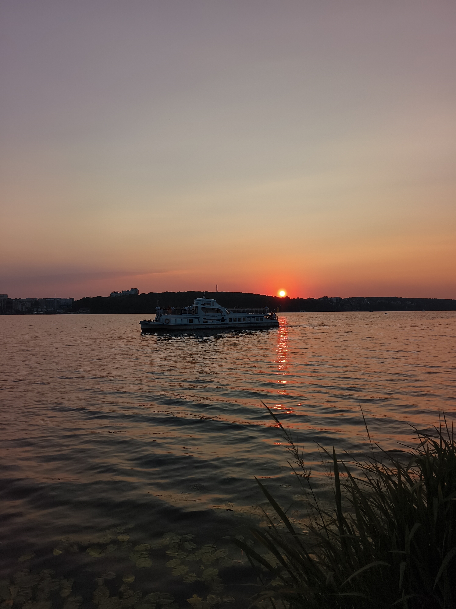 This is a photograph taken at sunset on Ternopil Lake in Ternopil, Ukraine. The sun is partially obscured by the horizon and appears as a bright orange disk, casting a reflection on the rippling water. A white passenger boat with several people visible on its decks is moving across the lake from left to right. In the background, a tree-covered shoreline is visible, with buildings and structures indicating an urban area. The sky displays a gradient of colors, transitioning from pale purple at the top to shades of orange and pink near the horizon. In the foreground, tall grasses and some water lily pads are partially submerged at the edge of the water. The overall lighting suggests a calm evening atmosphere.