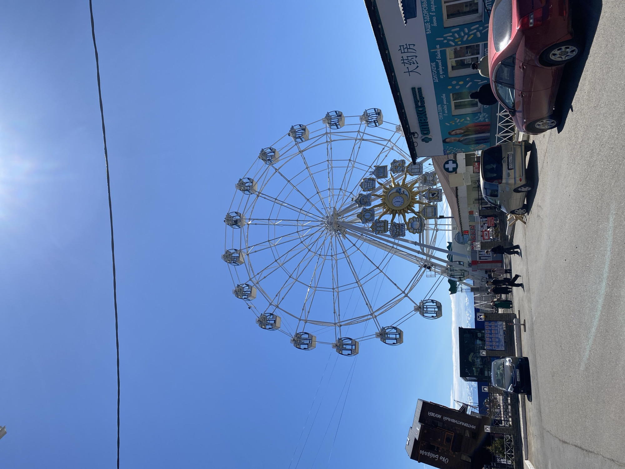 This image depicts a Ferris wheel in Listvyanka, Russia, set against a clear blue sky. The wheel has a white frame with enclosed gondolas, and at its center is a decorative sun-shaped design with a clock. Adjacent to the Ferris wheel is a building with signage in both Russian and Chinese, indicating a pharmacy ( аптека and 大药房).
Cars are parked along the side of the street, and pedestrians are walking nearby. The angle of the shot suggests that the image was taken from a low vantage point, emphasizing the height of the Ferris wheel. There are billboards, utility poles, and other urban features in the background, confirming this is an urban or tourist location in Listvyanka.