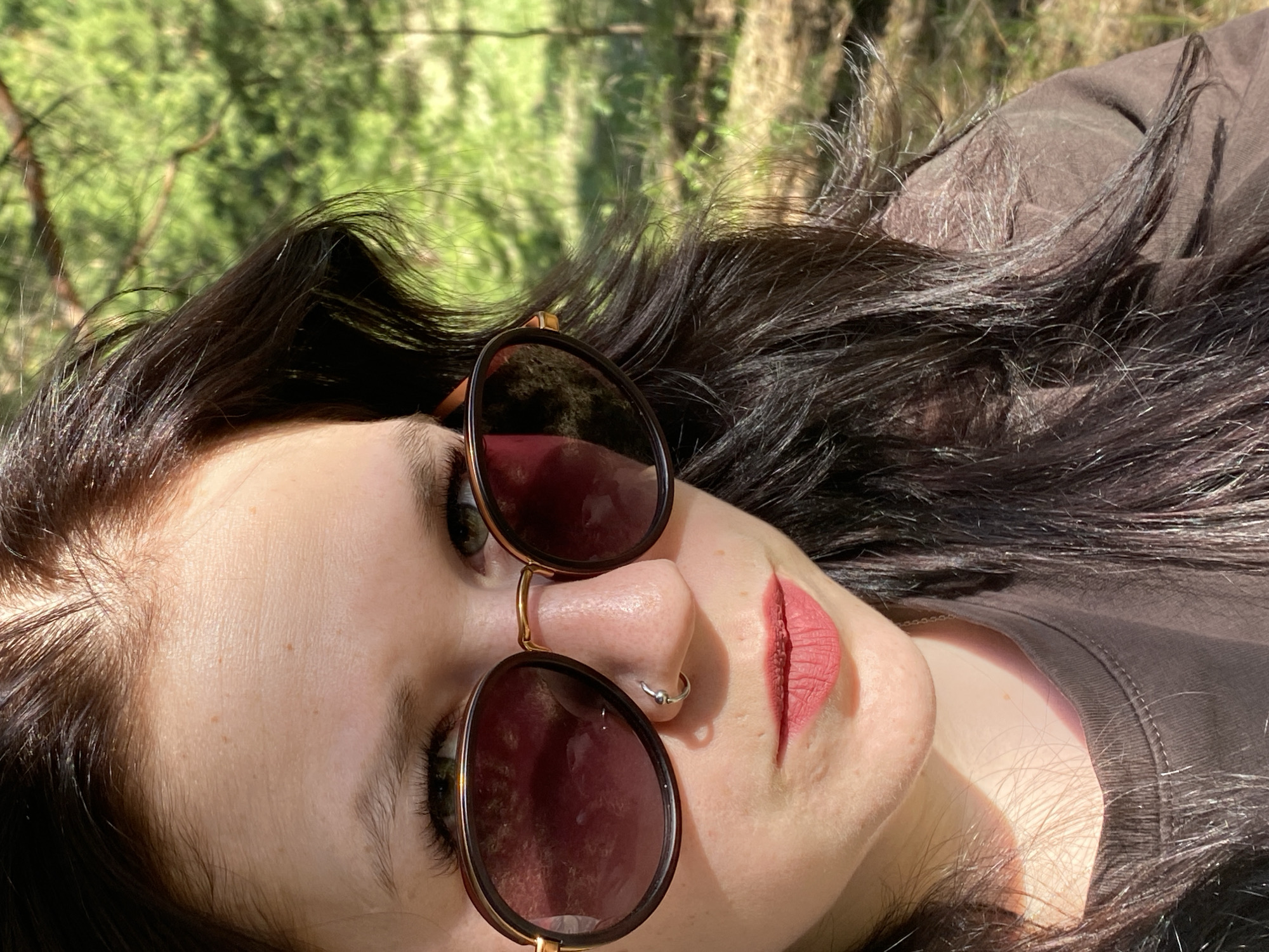 A woman is lying on her back in an outdoor setting, likely a park or wooded area in or near the city of Фоки, Russia. She is wearing sunglasses and a brown t-shirt. Her dark hair is spread around her head. Her skin is pale and freckled. She has a nose ring. The background shows blurred greenery and sunlight filtering through trees.