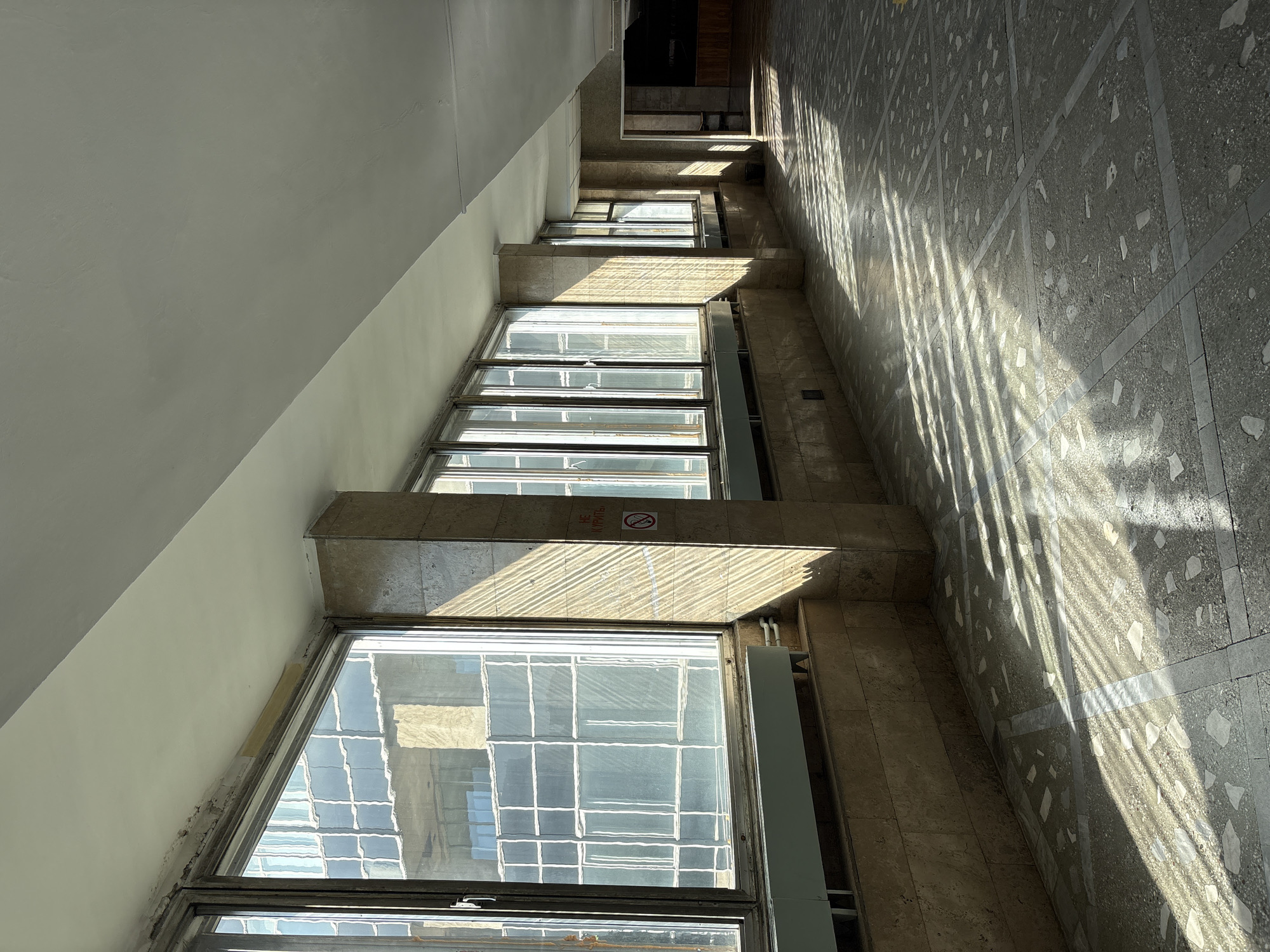 The image captures an architectural interior, looking downwards into a multi-story space, likely a stairwell or a window-lined corridor. On the left, a plain, light-colored wall angles sharply into the frame. To its right, a series of large, multi-paned windows are stacked vertically, extending into the depth of the image. These windows reflect grid-like patterns, possibly from an exterior facade or other structures.
The right side of the space features a tiled floor, likely terrazzo, with a distinct grid pattern and embedded lighter-colored stone fragments. Strong, angular sunlight streams into the space, casting prominent, striped shadows across the tiled floor and the concrete or stone ledges that separate the window sections. These long shadows suggest it is either morning or late afternoon on a sunny day.
A red circular "No Smoking" sign is visible on one of the ledges between the windows. The sign clearly shows a crossed-out cigarette and the Russian text "НЕ КУРИТЬ," which translates to "NO SMOKING." The building's construction, characterized by its robust materials and window design, suggests an older structure, possibly from the Soviet era, consistent with the location context of Izhevsk, Russia. There are minor signs of wear and age around the window frames and walls. No people or specific activities are depicted.