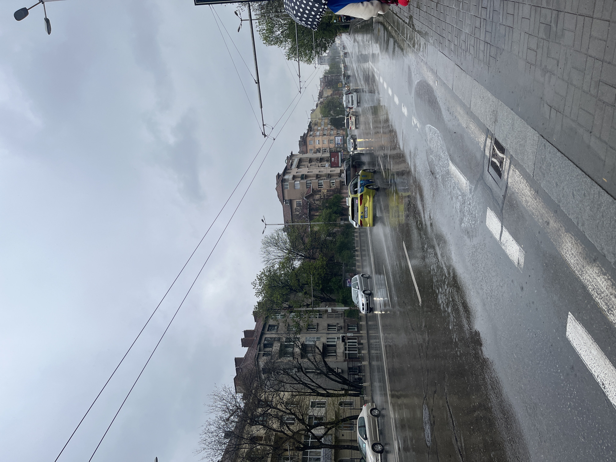 The image captures an overcast, rainy day on a city street in Sofia, Bulgaria. The street is wet with visible puddles and reflections, indicating recent or ongoing precipitation.
**Subjects:** The main subjects are cars, buildings, and a person partially visible under an umbrella. A bright yellow taxi with "OK SUPERTRANS" written on its side and license plate "C 9214 HT" is prominent in the foreground, driving away from the viewer. Other cars, including white and silver sedans, are also on the wet road. A pedestrian holding a black and white polka-dotted umbrella is seen on the right edge. The street is lined with multi-story residential or commercial buildings, exhibiting a typical Eastern European architectural style, some with balconies and air conditioning units. Trees, some bare-branched and some with green leaves, are interspersed along the sidewalk and in front of the buildings. Overhead wires, possibly for trolleybuses or trams, stretch across the scene.
**Activity or event taking place:** The scene depicts everyday urban life on a wet, rainy day. Cars are either driving or parked along the street. A person is walking, using an umbrella for protection from the rain.
**Notable details:** The time of day is daylight, characterized by a heavily overcast, cloudy sky that lends a muted, somewhat gloomy atmosphere to the scene. The pervasive wetness is a dominant detail, with reflections on the asphalt and visible puddles. White road markings, including dashed lines and solid lines, are present on the street. A storm drain grate is visible on the wet sidewalk.
**Visible text:** The most clearly visible text is "OK SUPERTRANS" on the side of the yellow taxi, along with its license plate "C 9214 HT". Another license plate, "CB 9811 AP", is partially visible on a white car further down the street.