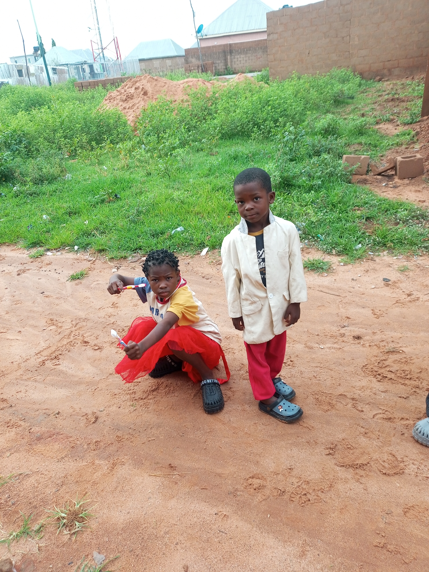 Two young children are in a sandy outdoor area. The child on the left is a girl with dark curly hair, wearing a yellow and blue t-shirt and a red ruffled skirt. She is crouched down, holding a toothbrush and a spoon. The child on the right is a boy wearing a light-colored jacket over a dark t-shirt and red pants. He is standing and looking towards the camera. The background shows green grass, a pile of sand, and some brick buildings under a bright sky, suggesting it is daytime. The overall setting appears to be a construction or undeveloped area.