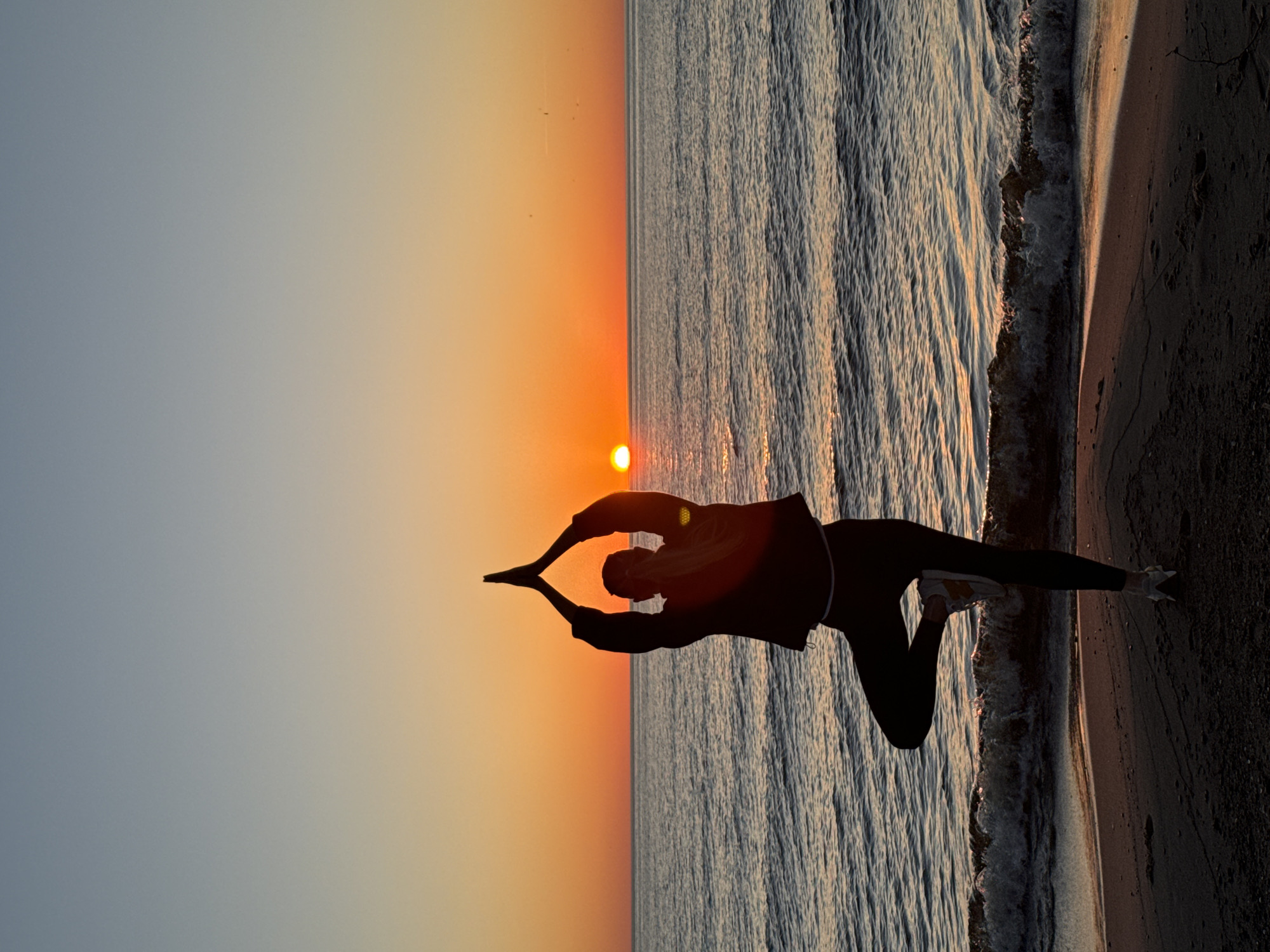 A silhouette of a person performing a yoga pose on a sandy beach with water in the background, possibly at sunset. The sky is orange and the water reflects the light. The photo appears to have been taken in Garciems, Latvia. The pose involves the person's hands meeting overhead, seemingly framing the sun.