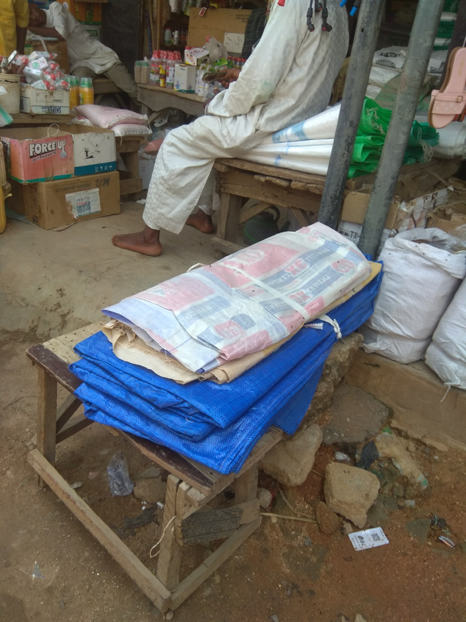 The image depicts an outdoor market stall in Jalingo, Nigeria. In the foreground, a wooden bench holds several stacked items, including a pile of blue tarps, a beige tarp, and a large, folded bundle wrapped in a white and pink woven material. The white and pink bundle is tied with white cord and has visible text and graphics that suggest it may contain bags of cement or similar goods. Behind the bench, a person wearing a white robe and trousers with patterned trim is partially visible, with their bare feet on the ground. They appear to be sitting or resting. To the left of the bench, a cardboard box labeled "FORCE UP GLYPHOSATE" is visible, stacked on top of another cardboard box. Other market goods, including sacks and bottles, are present in the background. The ground is dirt and concrete. The overall impression is a busy, utilitarian market environment.