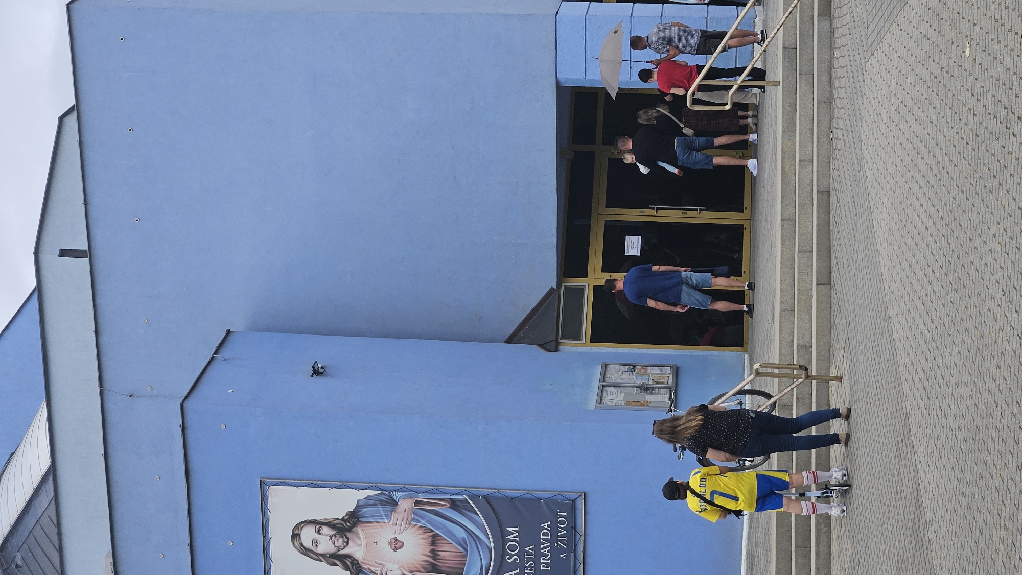 The image captures the exterior of a modern church building in Prešov, Slovakia, characterized by its striking bright blue facade. A large poster featuring an image of Jesus Christ is prominently displayed on the left side of the building, with Slovak text below it. In front of the entrance, a group of people are gathered. Two individuals, one a child on a scooter wearing a yellow jersey with "RONALDO" and the number "7" on it, and another person, stand near a bicycle. Further back, near the double glass doors of the entrance, several adults and a child are seen. Some are holding an umbrella, suggesting possible rain. The ground in front of the building is paved with interlocking gray bricks, and a set of wide concrete steps leads up to the entrance. The architecture of the church is angular, with layered sections of blue.