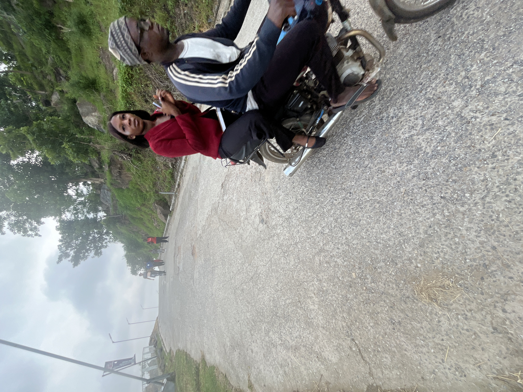 In Abuja, Nigeria, a scene unfolds on an asphalt road ascending a hill, bordered by lush green vegetation and rocky outcrops. The sky is overcast, suggesting either early morning or a cloudy day. In the foreground, a motorcycle carries two individuals. The rider, a male wearing a patterned cap and glasses, is seen from the side. The passenger, a female in a red jacket and black pants, is positioned behind him, holding a phone. Their body language indicates they are engaged in conversation or simply traveling together. Further up the road, a group of people are walking, some in athletic wear, suggesting a recreational activity like hiking or jogging. The overall impression is a blend of urban life and natural surroundings, characteristic of many areas in Abuja.