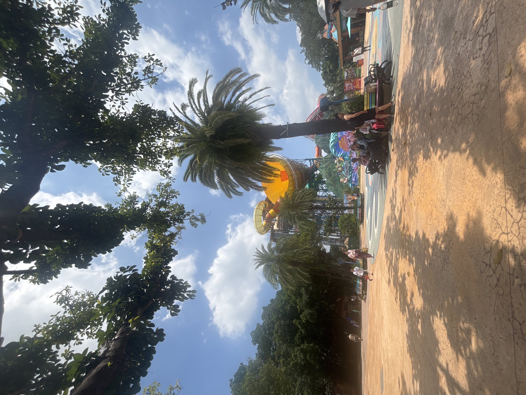 This image depicts an outdoor scene at a water park in Panyu District, China, on a sunny day with a blue sky and scattered clouds. In the foreground, an orange paved area is partially covered by the dappled shadows of overhead trees. Several people are visible, some walking and others seated on benches.
Dominating the middle ground are palm trees and a large water slide structure with a yellow and orange tube. The water slide complex appears to be a central attraction, with various platforms and support structures visible. To the left, a dense green forest forms the background.
Further in the background, to the right, there are buildings that appear to be part of the water park's amenities, including kiosks or food stalls, with colorful signage. The overall atmosphere suggests a recreational environment.