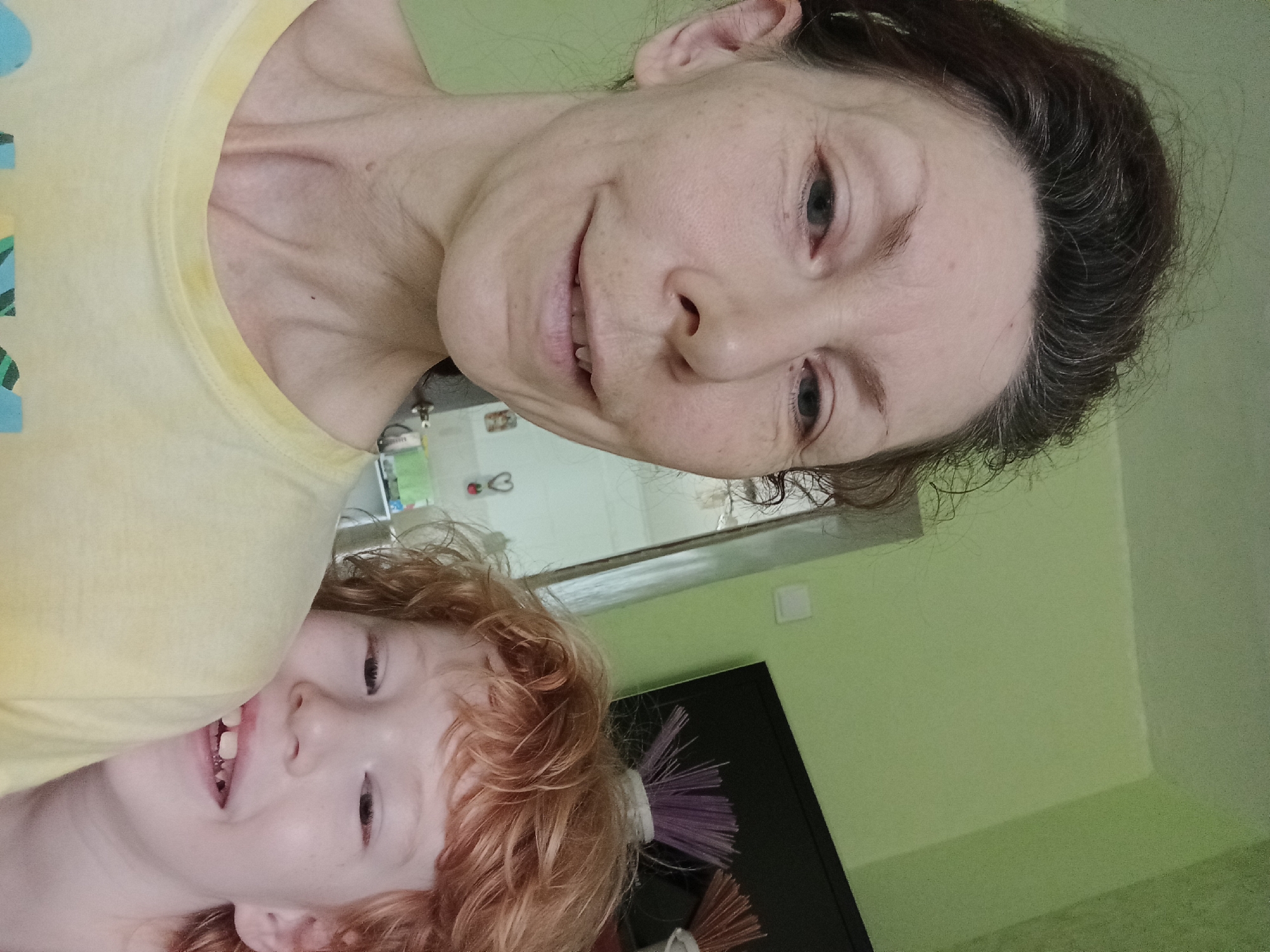 In a close-up selfie taken indoors, an adult with brown hair and a fair complexion is positioned on the left side of the frame, looking at the camera with a slight smile. To the right, a child with red, curly hair is visible, also looking towards the camera with a wide smile, teeth showing. The background consists of a light green wall and a black television screen on which a cluster of purple and orange items are placed. The adult is wearing a yellow t-shirt, and the child is partially obscured by the adult's shoulder. There are no discernible landmarks indicating the city of Blagoveshchensk, Russia.
