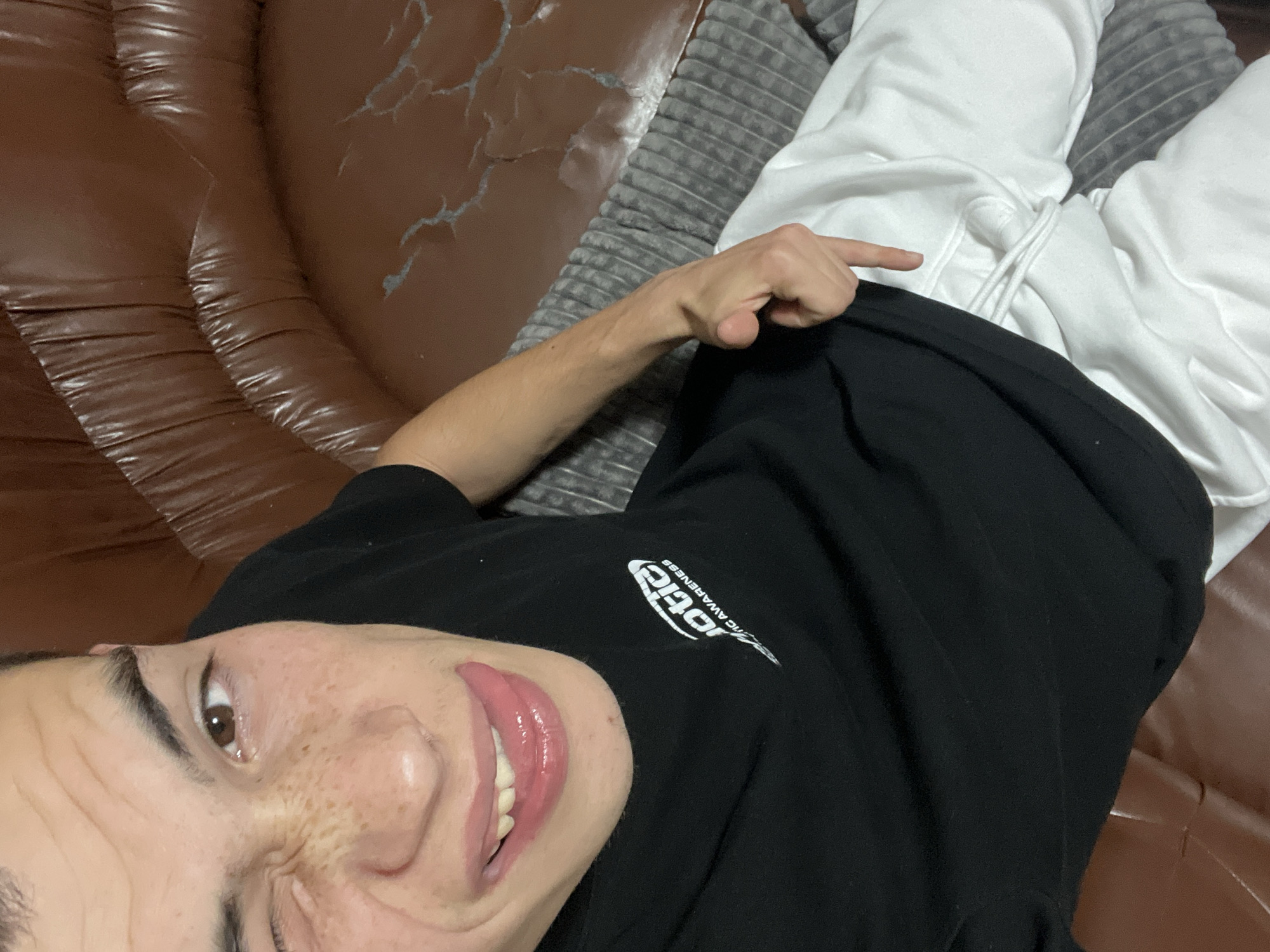 A person lies prone on a brown, possibly leather, couch. They are wearing white sweatpants and a black t-shirt with a white logo visible on the chest. Their right hand is raised, pointing a finger towards the waist area of the sweatpants. The couch appears to be worn, with visible cracking on the surface. A gray, textured pillow or cushion is also present on the couch. The image is shot from a slightly angled overhead perspective. There is no information available to suggest the image was taken in Plovdiv, Bulgaria.