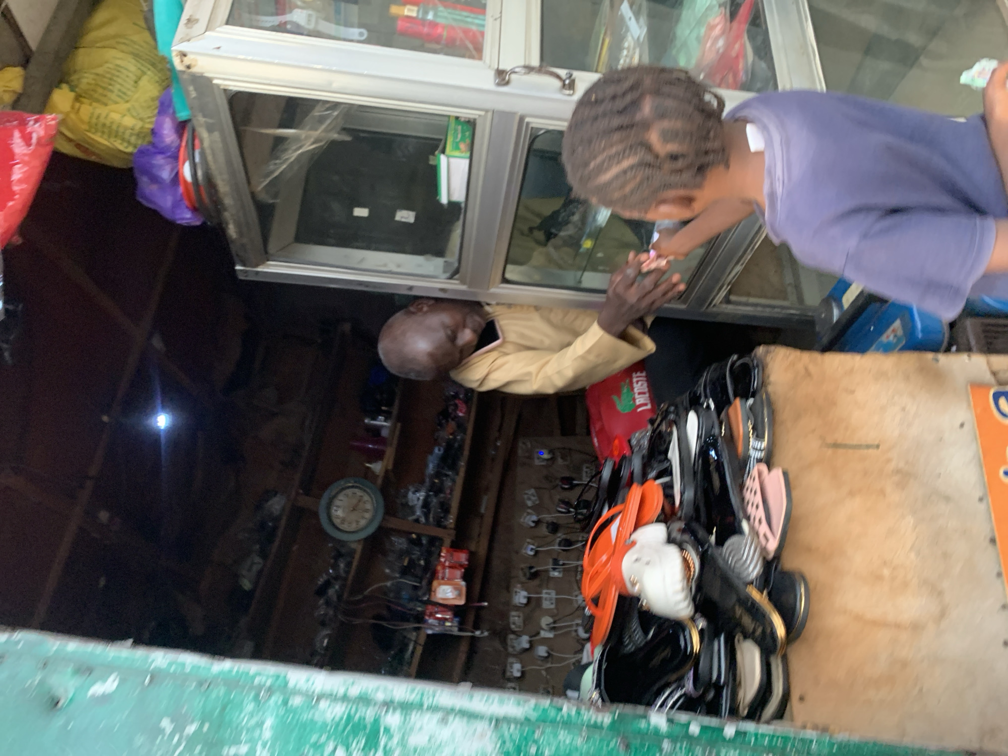The scene takes place at a small street-side business in Gura Top, Nigeria. A man with a bald head and wearing a tan shirt is situated inside the business, potentially a shop or kiosk, leaning towards the glass window. A younger person with braided hair, wearing a blue t-shirt, is standing outside the window, seemingly interacting with the man inside, possibly paying for something. A clock and various objects line the inside walls of the business. Shoes are stacked on a wooden surface on the right of the kiosk. Bags in the top left corner.