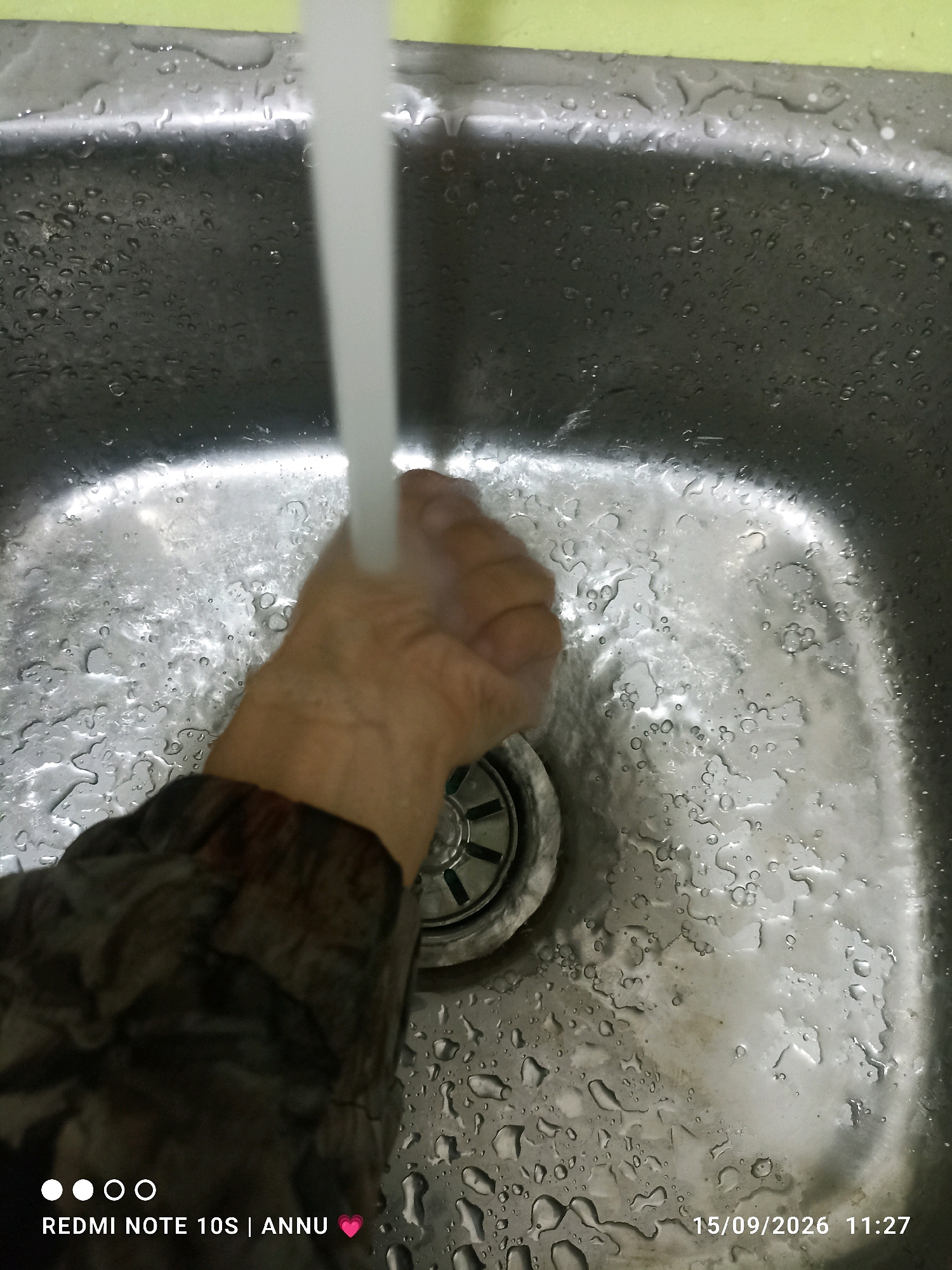 An overhead shot captures a hand being washed under a stream of water in a stainless steel sink. The sink is wet with numerous water droplets scattered across its surface. To the bottom left, a watermark displays "REDMI NOTE 10S | ANNU" along with a pink heart icon. To the bottom right, a date and time watermark reads "15/09/2026 11:27". The image does not provide any specific details or context that would allow for the identification of the location as Pampore, India.