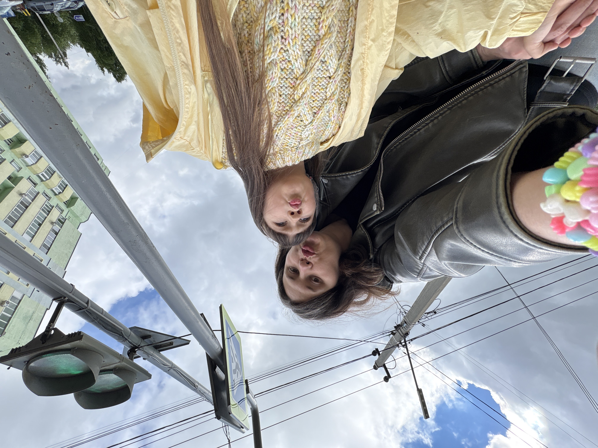 A low-angle shot, seemingly a selfie, captures two young women looking down into the camera with playful, pouty expressions. The image is taken from a street level, pointing upwards towards the subjects and the sky.
In the foreground, one woman has long brown hair and is wearing a dark (likely leather) jacket. Her arm is visible on the right, adorned with a colorful, multi-beaded bracelet. Behind her and slightly to the left, the second woman (or possibly the same woman's reflection/twin) has long, straight brown hair. She is wearing a light yellow cable-knit sweater, layered under a pale yellow jacket.
The background reveals an urban environment under a partly cloudy sky, with patches of blue visible amidst the white and grey clouds. To the left, a multi-story apartment building with a distinctive green and pale yellow facade, numerous windows, and external air conditioning units is visible. In front of the building, a grey pole holds a green-lit traffic signal and a blue-and-white pedestrian crossing sign. Utility poles and a web of overhead wires crisscross the sky on the right side of the frame. Bare trees are visible in the far background, with a car parked on what appears to be a street. A blurry sign with the possible text "BATA" can be faintly discerned in the distant background. The scene depicts a daytime setting with an overcast but bright sky, conveying a casual and lighthearted mood.