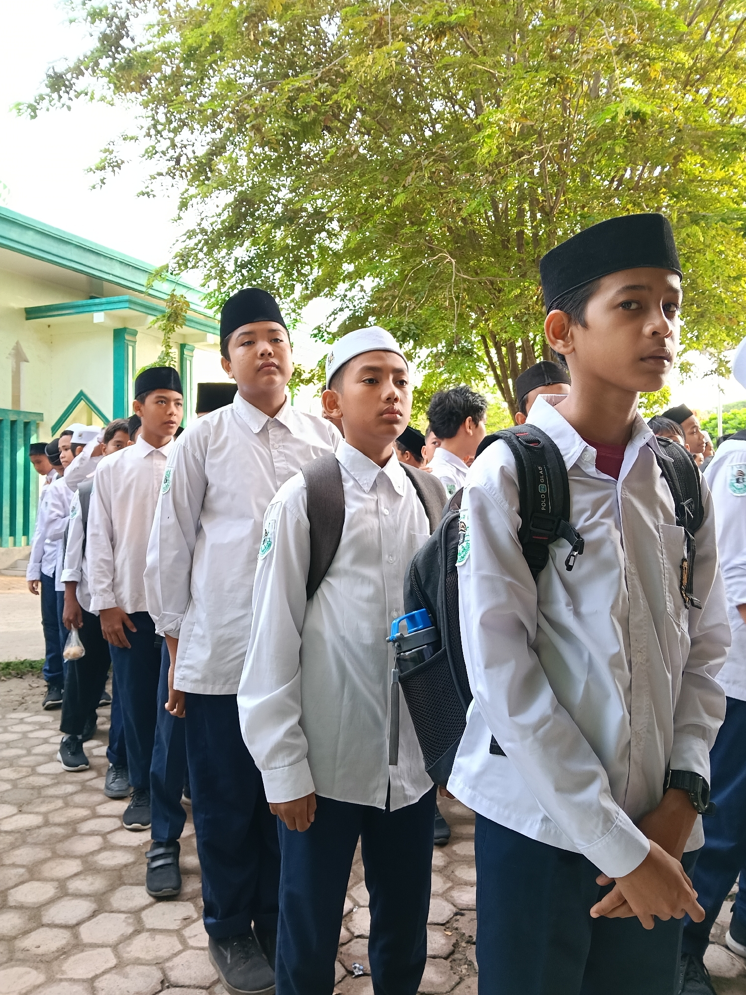 A group of young male students are standing in a line outside a building. They are wearing white shirts and dark pants, and some are wearing black or white caps. Some students have backpacks. The building behind them is light-colored with a green trim. A large tree is visible in the background. The scene appears to be in Lhokseumawe, Indonesia, as indicated by the clothing and setting.